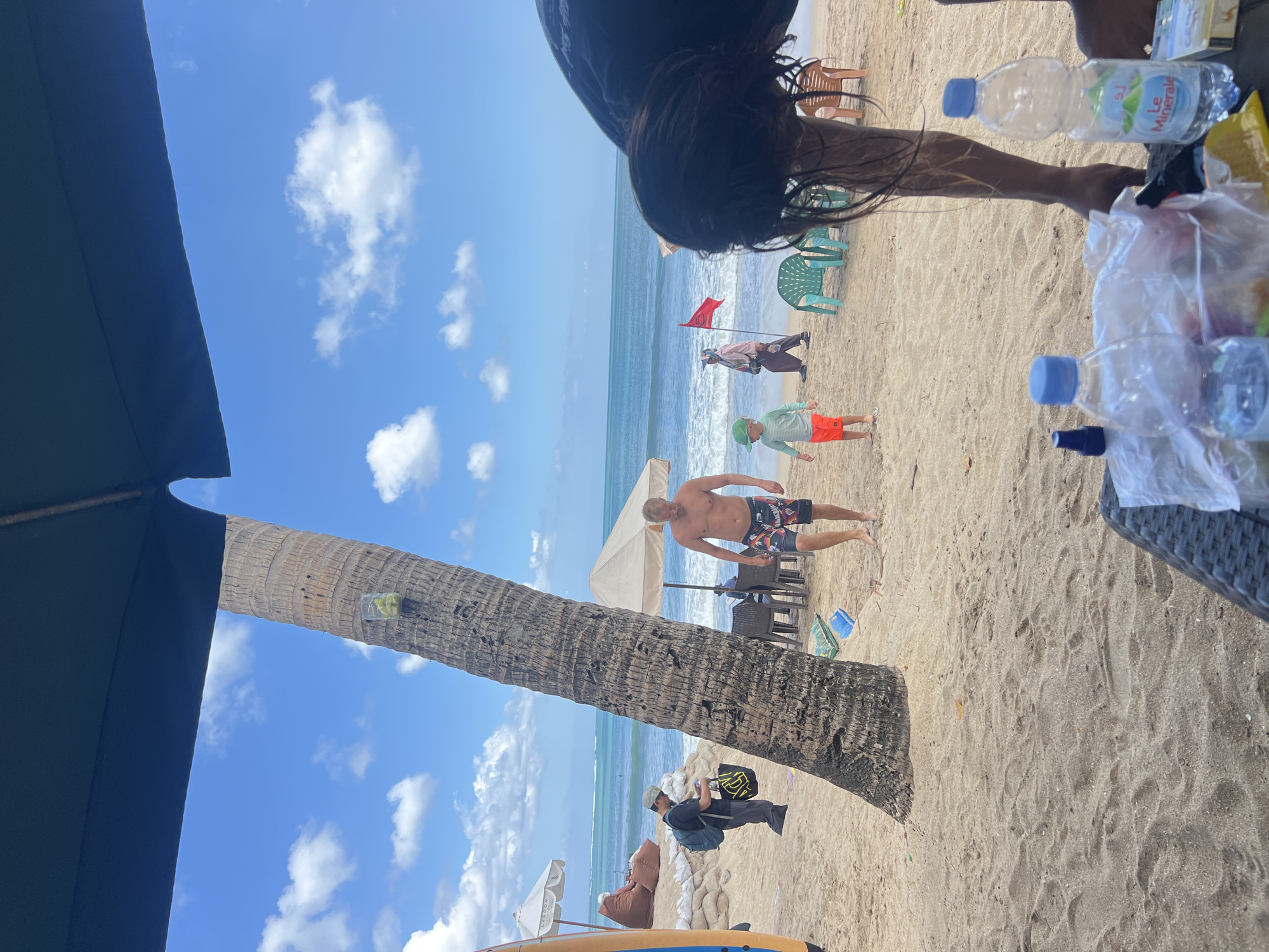 The image depicts a beach scene in Kuta, Indonesia, under a bright, partly cloudy sky. A large palm tree dominates the foreground, its trunk textured and weathered. In the middle ground, a shirtless man with short blond hair walks towards the viewer on the sandy beach, wearing patterned swim trunks. To his left, a young child in a light blue shirt and orange shorts is walking away from the viewer, towards the ocean. Further back, a person wearing a headscarf and a light-colored top walks, with a red flag planted in the sand nearby. Beach chairs and umbrellas are scattered across the sand. In the foreground, a table is set up with plastic bottles of water and bags, suggesting a resting spot. The ocean is visible in the background with gentle waves breaking on the shore.