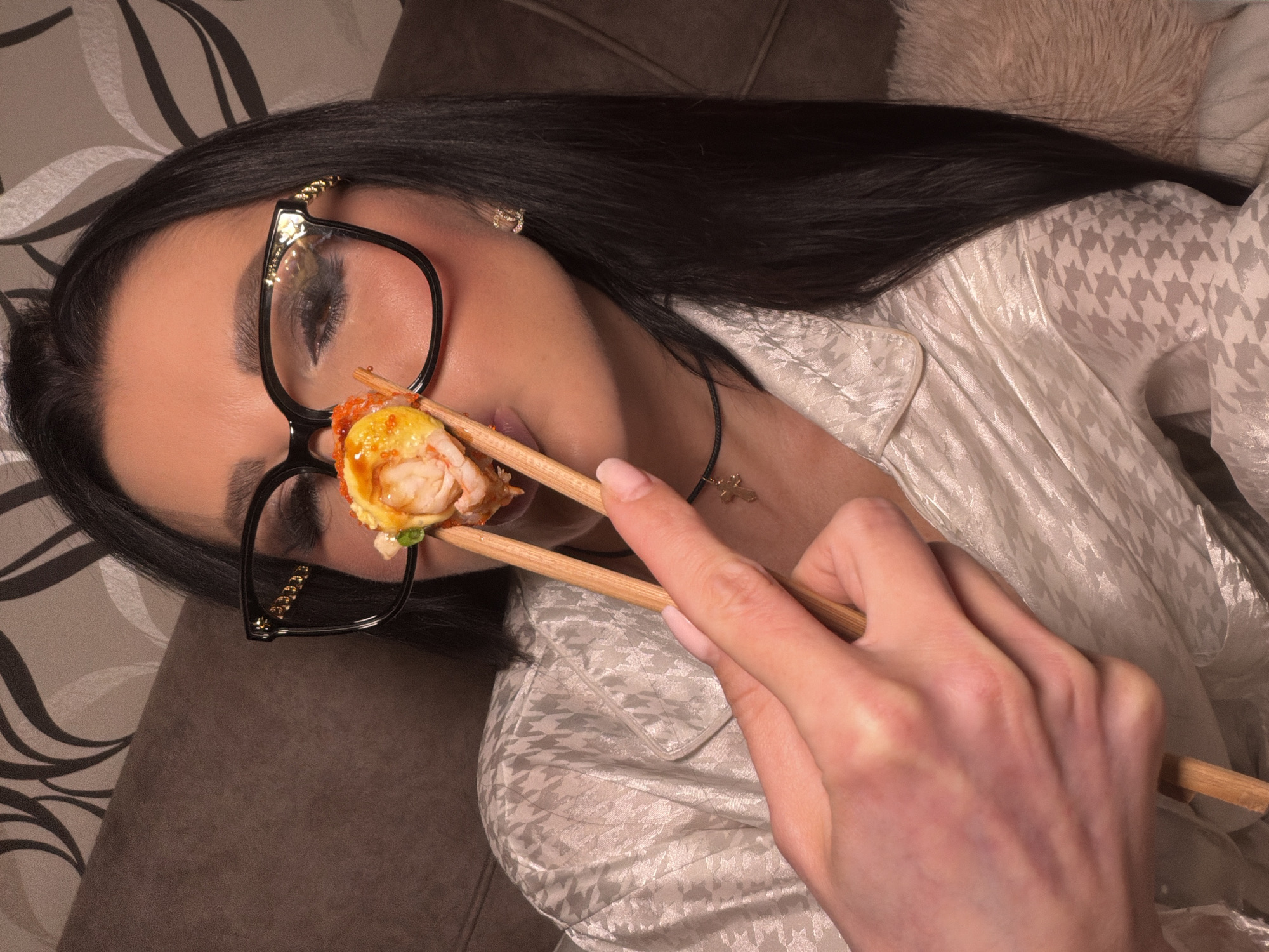 A woman with dark hair and glasses lies on a sofa while holding a piece of sushi with chopsticks. She wears a patterned white shirt and a necklace with a cross pendant. The sofa has a patterned cushion and a brown surface. The image appears to be taken in an indoor setting. No information in the image confirms the location in Svkh Voronezhskiy, Russia.