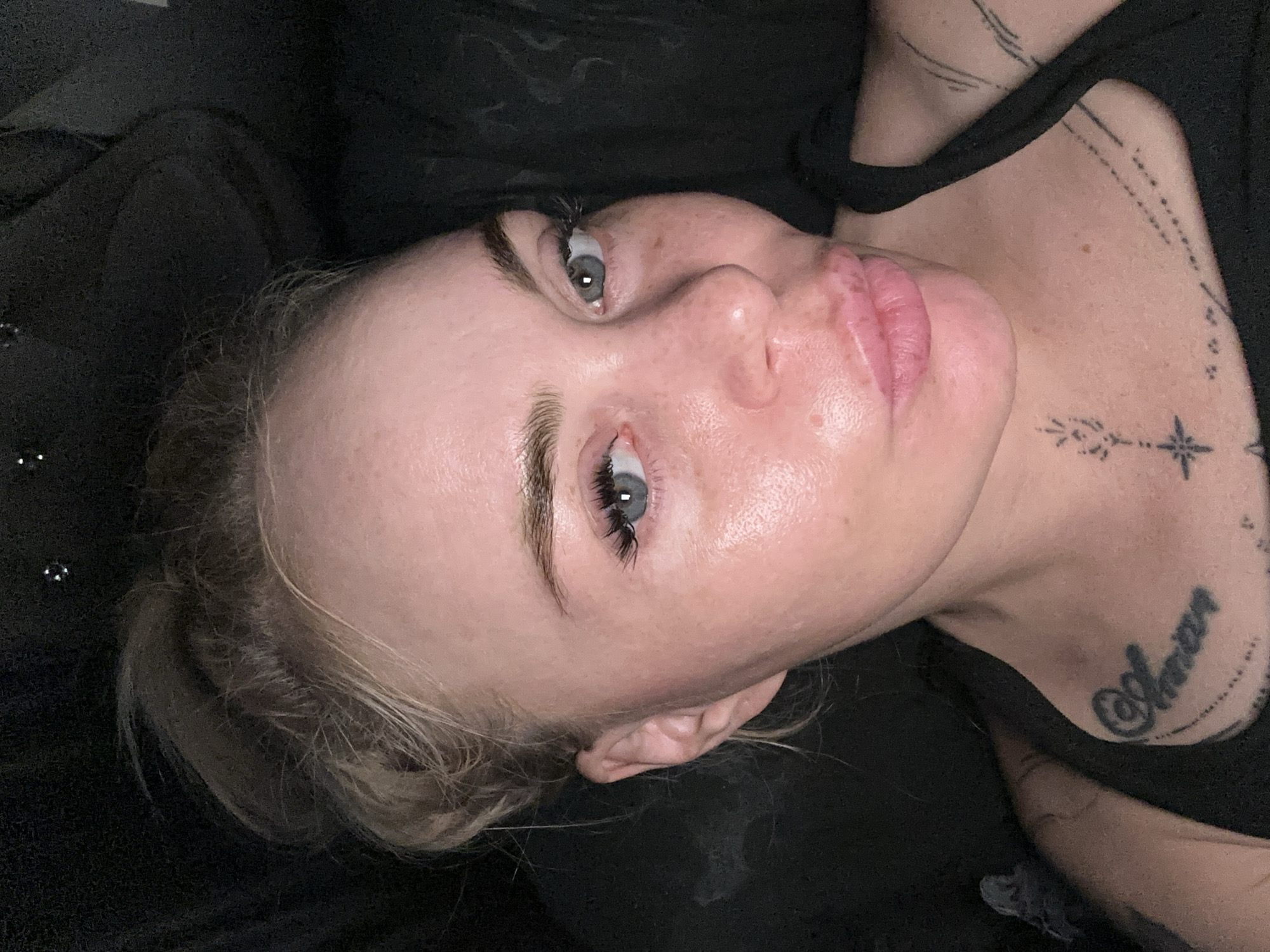 A woman with blonde hair pulled up in a messy bun is looking at the camera, pouting her lips. Her eyes are open and her gaze is direct. She has on minimal makeup, with some noticeable blush and mascara. There are tattoos visible on her neck and chest. The background is dark and appears to be indoors, possibly a couch or a dark room.