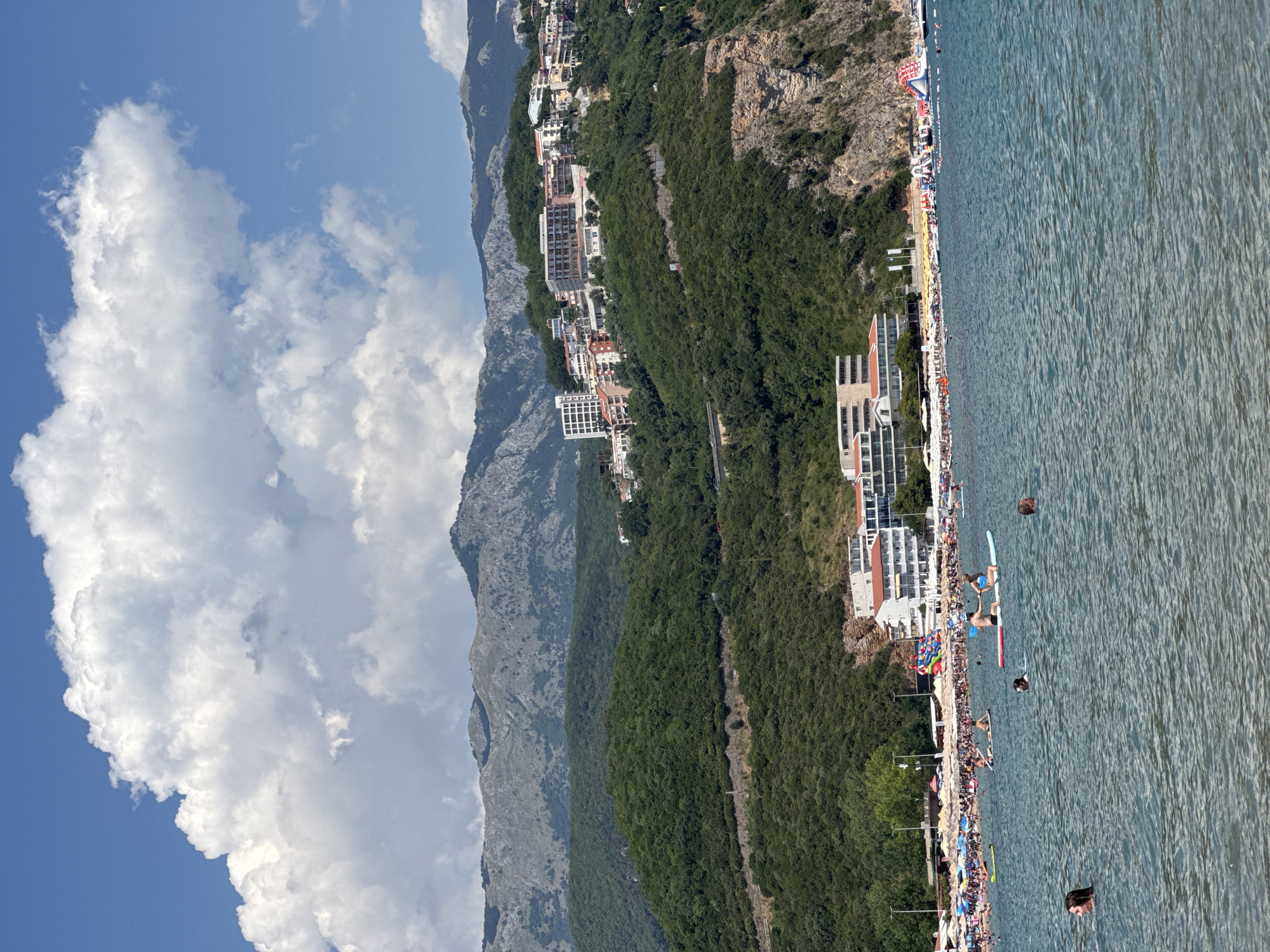 A scenic view of the coastline near Prijevor, Montenegro, showcases a vibrant beach scene against a backdrop of lush green hills and majestic mountains. The turquoise waters of the Adriatic Sea are populated with various people enjoying leisure activities. Several individuals are swimming, while others are on paddleboards, some in pairs, navigating the calm waters.
The beach itself is a lively hub of activity, packed with sunbathers lounging under numerous colorful umbrellas. Beach chairs are arranged in neat rows, indicating a well-organized resort. Along the shore, there's an inflatable water park, adding to the recreational offerings. In the distance, hotels and residential buildings are nestled into the verdant hills, their architecture reflecting a modern coastal style. The scene is further enhanced by the dramatic mountainous landscape that stretches across the horizon, with layers of peaks gradually fading into the sky, capped by a bright blue sky adorned with fluffy white clouds.