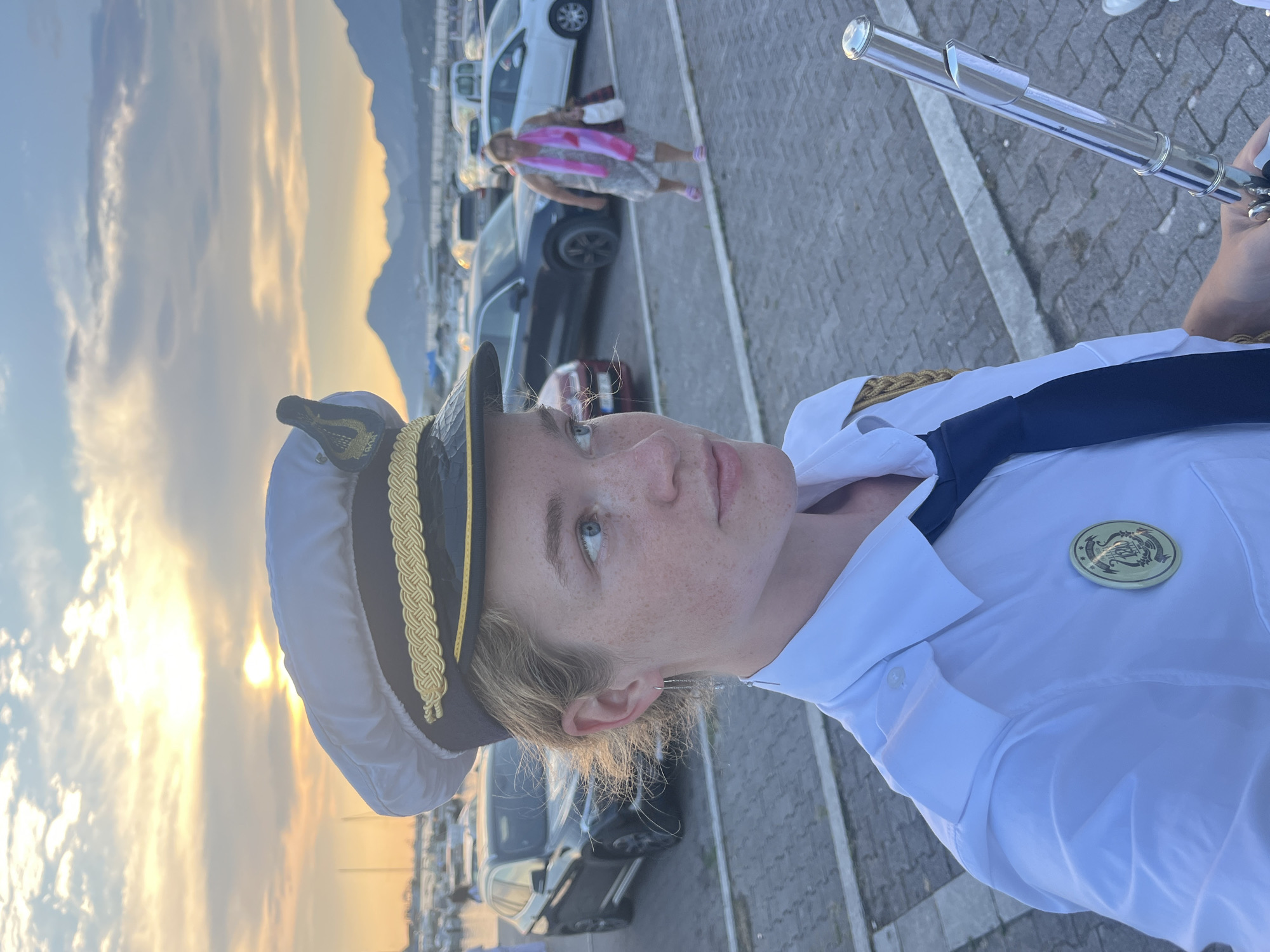 A young person in a white captain's uniform and hat poses for a photo with a flute. The sun is setting behind them, casting a warm glow on the clouds and mountains in the distance. The scene appears to be a parking lot next to a body of water, possibly a marina, with several cars and a boat visible in the background. The person has light skin and freckles and is looking off to the side with a slight smile. They are holding a silver flute horizontally in front of them. The uniform includes a dark blue tie and a gold emblem on the hat. The overall mood is peaceful and serene, with a hint of playfulness from the person's attire and the flute. The time of day is sunset, and the weather appears to be clear. The location is identified as Šušanj, Montenegro. There is no visible text in the image.