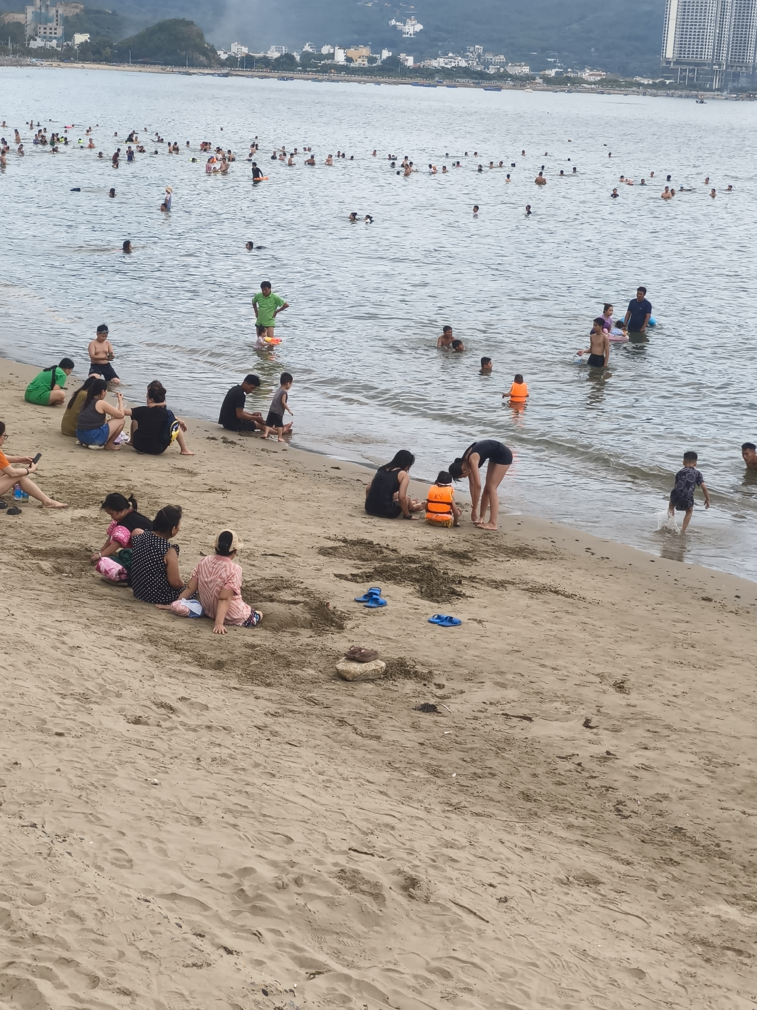 This media file depicts a crowded beach scene in Bac Nha Trang Ward, Vietnam. The primary subjects are numerous people, including adults and children, engaged in various activities. Some individuals are wading or swimming in the ocean, while others are sitting or playing on the sandy shore. Several children are wearing life vests, suggesting a family-friendly environment. Objects visible include beach towels, sandals, and a small bucket and spade.
The activity taking place is a day at the beach, with people enjoying the water and the sand. The weather appears to be overcast, with a hazy sky. The presence of a hilly landscape with buildings in the background and boats on the water indicates a coastal urban setting. The overall mood appears relaxed and recreational, typical of a beach outing. There is no visible text in the image.
