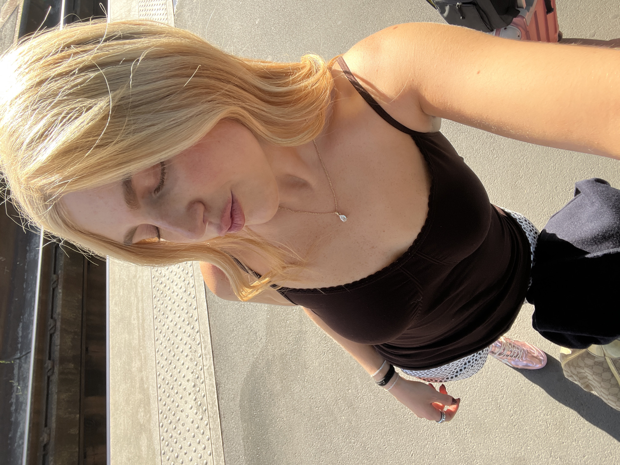 A medium-shot, high-angle, eye-level view of a light-skinned person, presumably female, with blonde hair, wearing a brown tank top, a gold necklace, and bracelets, standing on what appears to be a train platform in Paris, France. The platform has a textured, gray surface with tactile ground surface indicators. A metal rail runs along the edge of the platform. In the background, there are luggage pieces, including a pink suitcase. The person is holding the camera at arm's length, facing downwards at an angle. The person is wearing a skirt or shorts with a white and black polka dot pattern. Pink sneakers are also visible. A dark jacket is draped over a bag that is on the ground. The light suggests it is either morning or late afternoon.