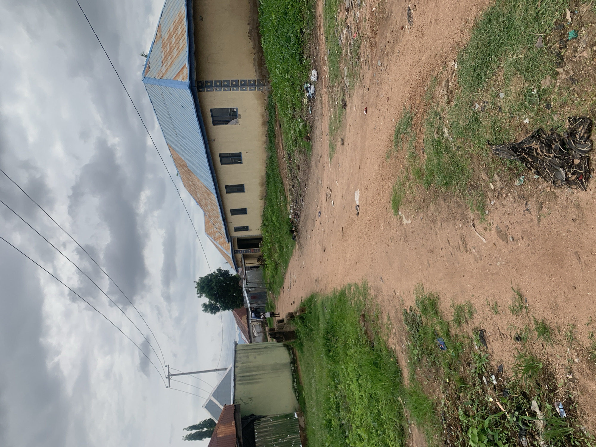 An outdoor medium shot shows a dirt road in a residential area in Jalingo, Nigeria, under a cloudy sky. The road is reddish-brown and has tire tracks visible. To the left of the road, there is a patch of green grass, followed by a light green building with a corrugated metal roof and fence. Further down the road, there is a person in a white shirt and dark shorts walking away from the camera. To the right of the road is a long, tan building with a blue and rust-colored corrugated metal roof. Several windows with dark frames are visible on the building. The ground next to the road on the right is also covered in grass and debris, including plastic bags and other refuse. In the foreground on the right side of the road, there is a pile of dark, textured material that appears to be fabric or possibly dried leaves. Electrical wires are strung across the sky, supported by a utility pole on the left side of the frame. The scene suggests a simple, lived-in environment.