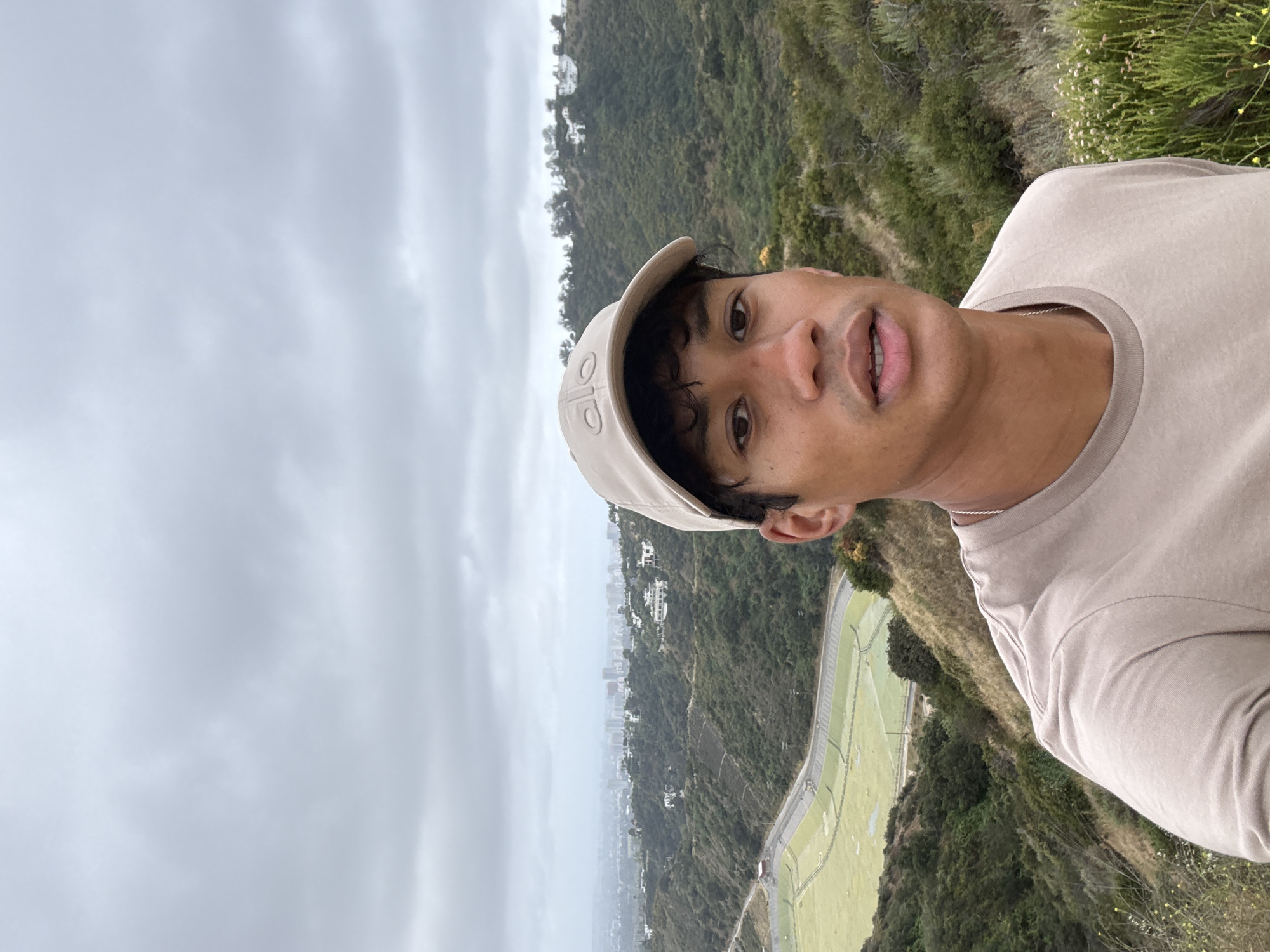 The image is a selfie taken by a man wearing a beige cap and matching T-shirt. He is positioned on a hillside overlooking the city of Los Angeles. The sky is overcast. In the background, a green, grassy area bordered by a concrete structure is visible. Further back, the cityscape is obscured by the overcast conditions. The surrounding terrain is a mix of greenery and trees.