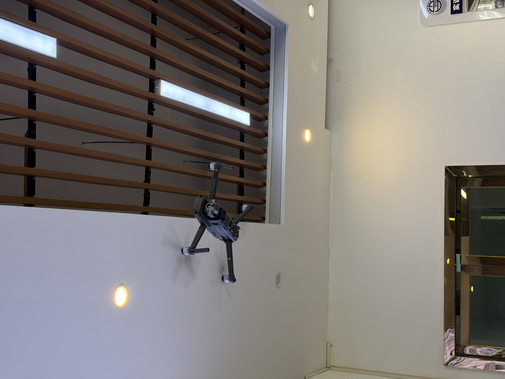 A drone hovers mid-air below a modern ceiling in what appears to be an indoor commercial space. The ceiling features a section with horizontal wooden slats, interspersed with modern rectangular lights. Below this, a plain white ceiling has recessed circular lights. The drone is a dark grey quadcopter with its arms extended and propellers attached, oriented downwards and slightly to the right. The scene is likely from the city of Jiangjin, China, given the context of the request. No people are visible in the image.