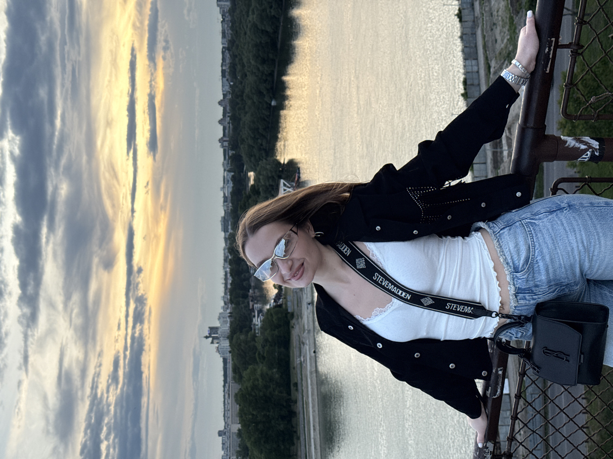A young woman stands on a bridge overlooking the Sava River in Belgrade, Serbia. She is leaning against the railing of the Branko's Bridge. The woman is wearing jeans, a white top, a black jacket, and sunglasses, and carrying a black purse with a Steve Madden shoulder strap. The time of day appears to be near sunset, with the sky displaying a mix of orange, yellow, and gray hues. Trees and buildings line the banks of the river.