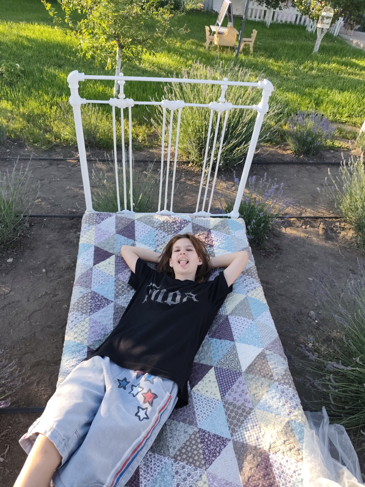 A young person with brown hair lies on their back on a quilted mattress on the ground. The person is wearing a black t-shirt with a graphic print and light blue shorts with red and white star designs and red and blue stripes along the sides. Their arms are behind their head, and their tongue is sticking out. The mattress is placed on the ground, possibly in a garden or park setting, with a white metal bed frame behind it. In the background, there is green grass, lavender bushes, and a white picket fence. The scene appears to be outdoors during daylight. The location is identified as Nechaiane, Ukraine.