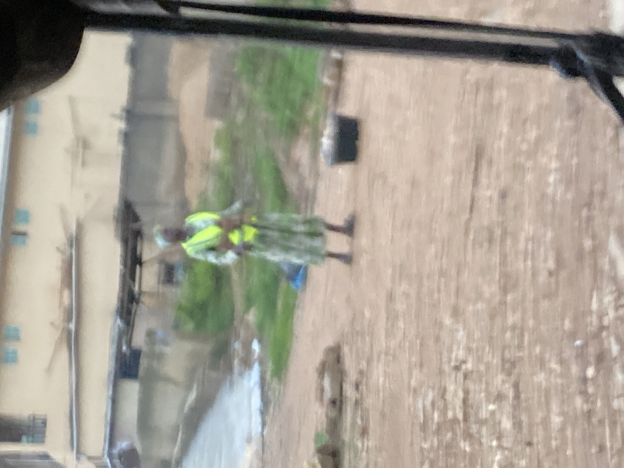 A blurred, medium-shot image depicts an outdoor scene on a dirt path, likely in Gwong, Nigeria. In the middle ground, a person wearing a long, patterned dress and a high-visibility yellow vest stands. The person is facing slightly to the left and appears to be holding something in their hands, though it is not clearly discernible. To the right of the person, a dark, round bucket filled with light-colored items sits on the ground. The background shows buildings with windows and what appears to be a corrugated metal roof structure. The ground is a mix of dirt, small stones, and patches of green grass. The foreground is dominated by a dark, out-of-focus object that obstructs a portion of the lower right side of the frame, possibly part of a vehicle or structure. The overall lighting suggests an overcast day.