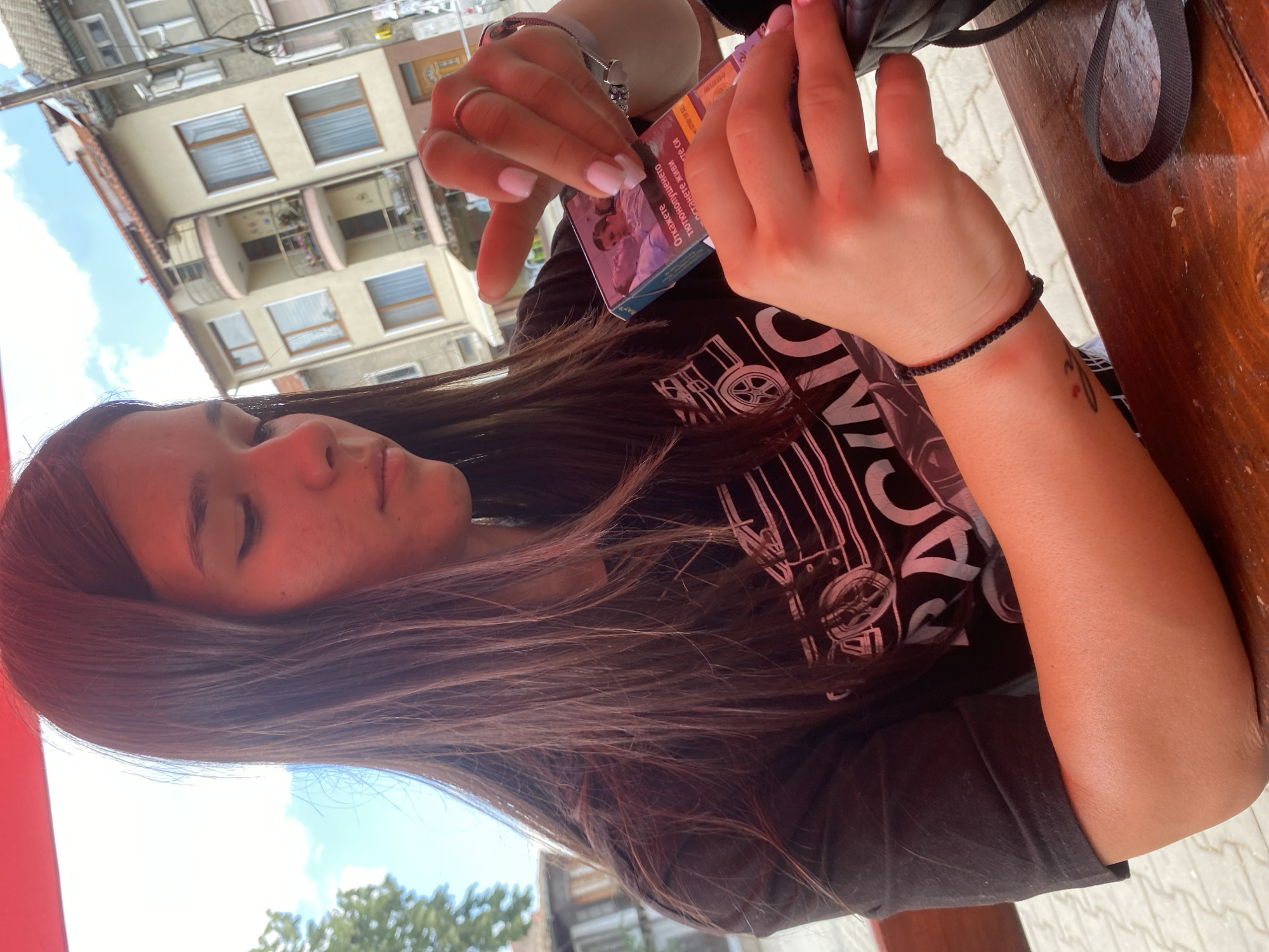 A young woman with long, dark brown hair is holding a pack of cigarettes in her hands, which are resting on a wooden table. She is wearing a black t-shirt with a white graphic design. Her fingernails are painted white, and she is wearing a silver ring on her left ring finger and a black beaded bracelet on her left wrist. The background shows buildings with balconies and windows, suggesting an urban or town setting, possibly in Sevlievo, Bulgaria, given the context. The sky is visible and appears to be blue with some clouds. The overall scene is casual, with the woman appearing to be engaged with the cigarette pack.