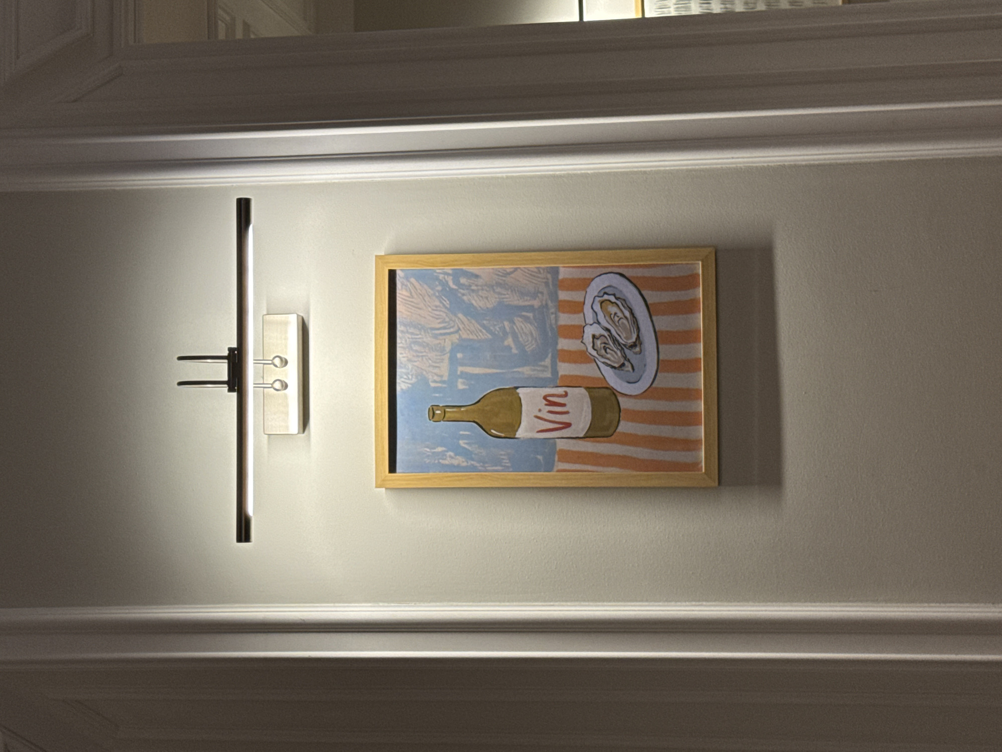 A framed picture hangs on a wall illuminated by a picture light above it. The picture depicts a wine bottle labeled "Vin" and a plate of oysters set against a background with a blue sky and orange stripes. The wall is painted a light neutral color and framed by decorative molding at the top and bottom of the image. The location is not discernible from the image.