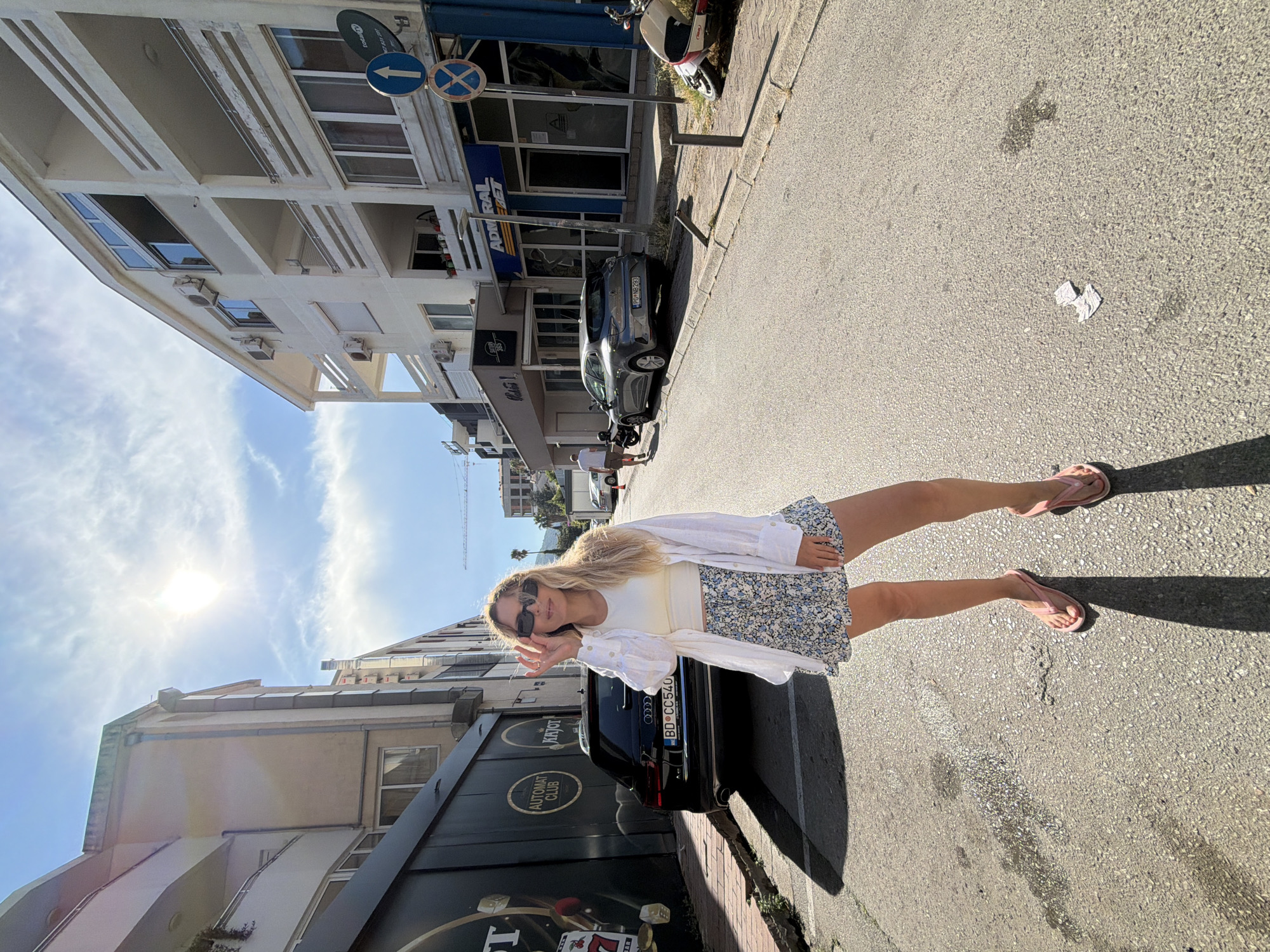 The image depicts a young woman posing outdoors on a sunny day. She has blonde hair and is wearing a white top, a white unbuttoned shirt, and a floral skirt. She is also wearing sunglasses and pink flip-flops. Her left hand is raised, and she is looking towards the camera with a slight smile.
The woman is standing next to a black Audi car parked on a street. Behind her, there are buildings lining the street, some with balconies and air conditioning units. The architecture suggests an urban setting, likely in a Mediterranean climate given the sunshine and clear sky. In the background, a construction crane is visible, indicating ongoing development.
The ground is paved asphalt, and there are some white paper tissues scattered on the street, along with a wet patch. The sunlight casts shadows on the ground, suggesting it is mid-day or early afternoon. There is some visible text on the building behind the car which includes "AUTOMAT CLUB" and "KRYOT", and a license plate on the car reads "BD CC 540". The overall atmosphere is casual and relaxed.