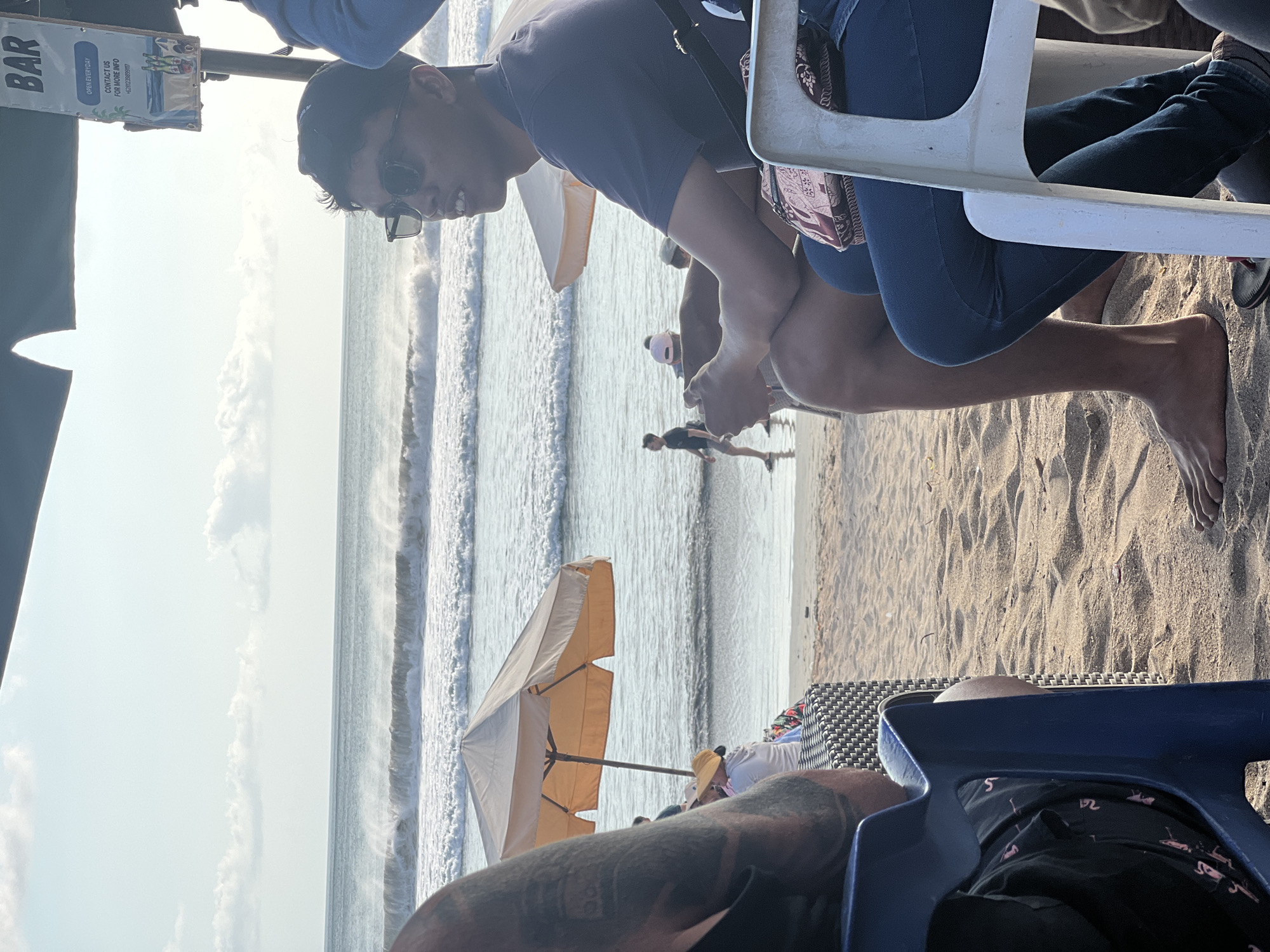 In Kuta, Indonesia, on a sandy beach with the ocean in the background, two people are seated in outdoor chairs. The person on the left is partially visible from the shoulder down, with a tattooed arm. The person on the right is a young man wearing sunglasses and a dark blue polo shirt. He is looking towards the right side of the frame and has his hands clasped. Behind them, a beach umbrella shades a table with other people. A sign for a "BAR" is visible hanging overhead. The ocean has moderate waves and the sky is partly cloudy.