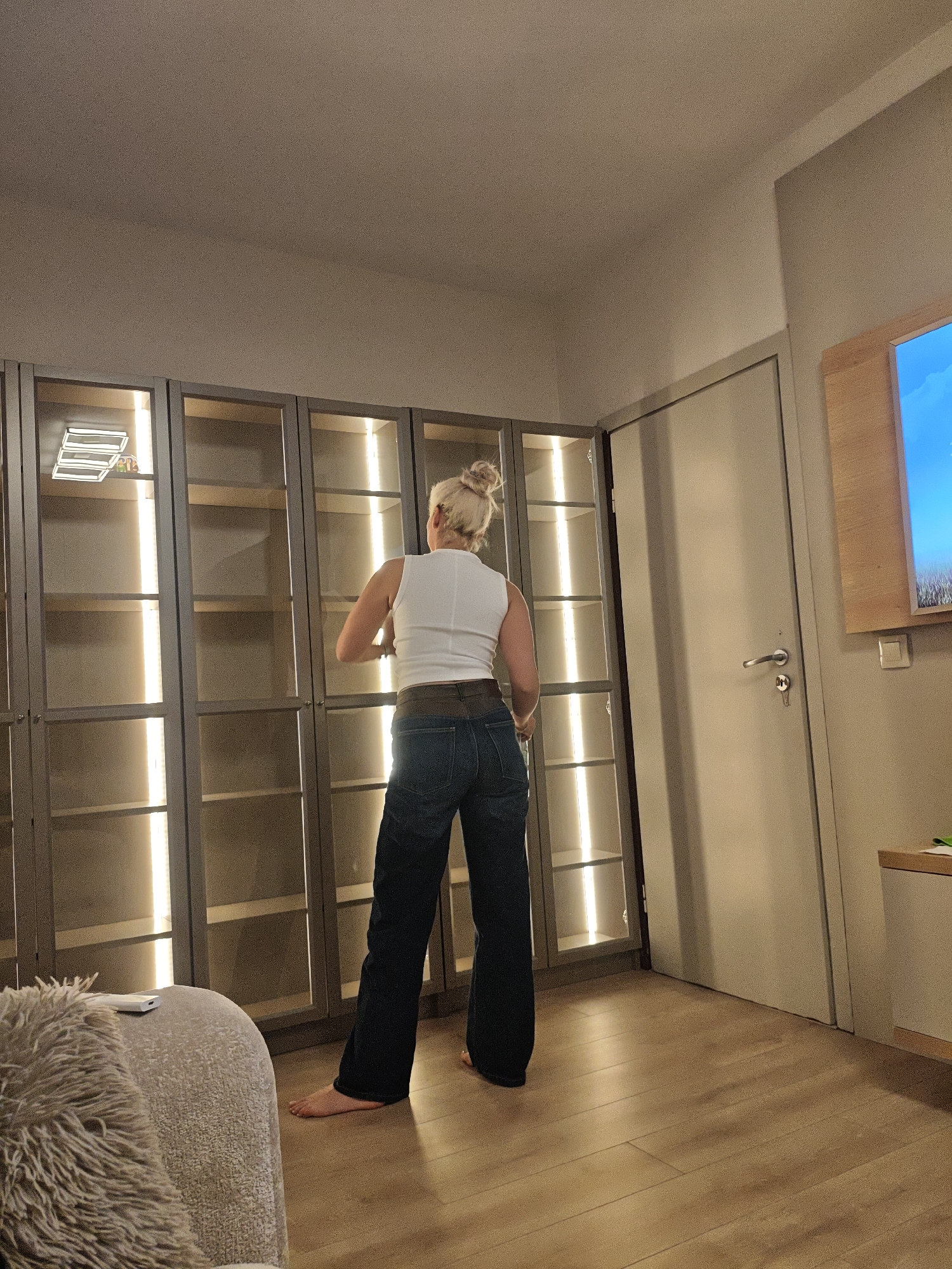 A full, medium-shot indoor photograph depicts a woman from the back, standing in front of a large, illuminated display cabinet. The location appears to be a residential interior, likely a living room or a similar living space in the city of Unknown, Unknown.
The woman is dressed in a white sleeveless top and dark blue, flared jeans, her hair is styled in a bun. She is barefoot. She appears to be either inspecting or arranging items within the display cabinet, which features multiple compartments with vertical lights.
To the right of the cabinet is a closed, plain door. Further to the right of the door, there is a television mounted on the wall, its screen showing a blue sky, and a partial view of the side table is also visible. In the left foreground, a corner of a plush, upholstered chair or sofa is visible. The lighting is warm, with a combination of artificial light from the cabinet and ambient light filtering through the room. The floor is covered with wooden planks, with their direction going away from the camera. The overall atmosphere suggests a cozy, domestic setting.