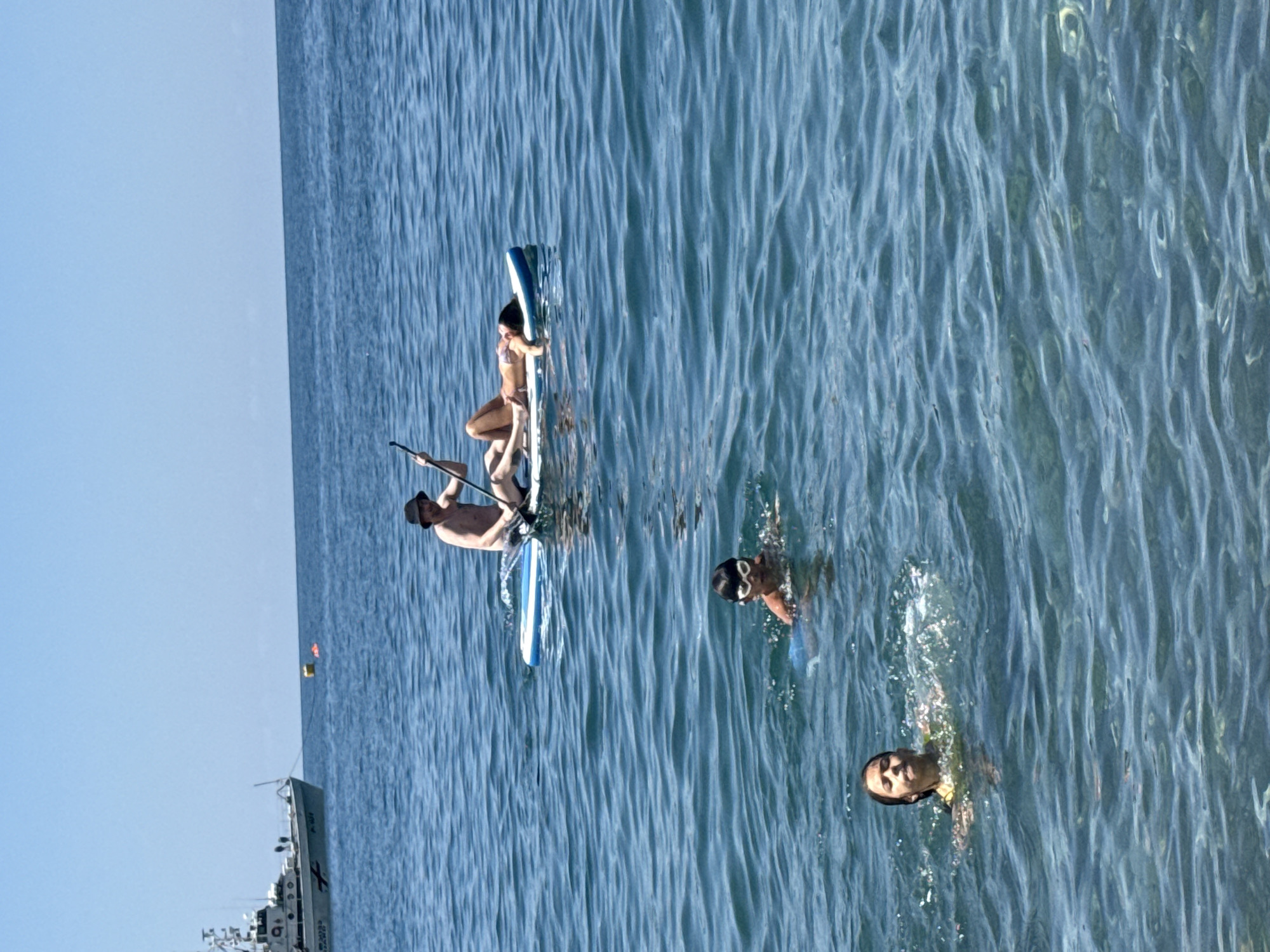 In the city of Sarpi, Georgia, a group of people are enjoying a day at sea. Two children are swimming in the clear blue water, with one child wearing goggles. In the background, a man is paddleboarding, and a woman is lying on the paddleboard. A boat is anchored in the distance, adding to the serene coastal atmosphere.