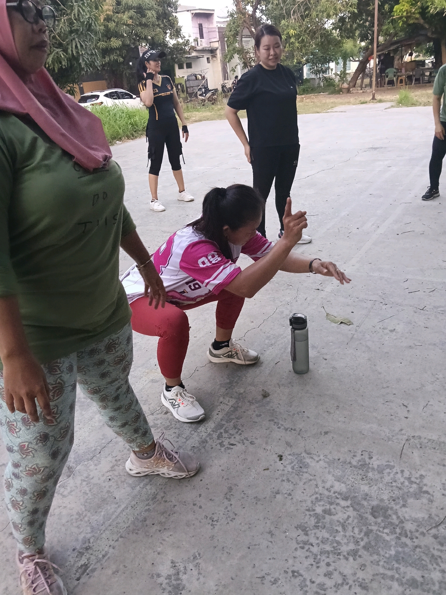A group of five women are exercising outdoors on a concrete surface in Jatimulya, Indonesia. In the foreground, a woman on the left wears a pink hijab and a green t-shirt with the words "DO THIS" written on it. She is also wearing patterned leggings and pink sneakers. To her right, a woman in a pink and white jersey with red leggings and white sneakers is performing a squat. Next to her, a water bottle stands upright on the ground. Further back, a woman in a black t-shirt and black pants stands with her hands on her hips. Another woman, wearing a dark green and yellow athletic shirt, black shorts with zippers, and a black cap with "SPORTS" written on it, stands with her hand on her neck. The background shows trees, a white car, and buildings. The overall lighting suggests it is daytime.