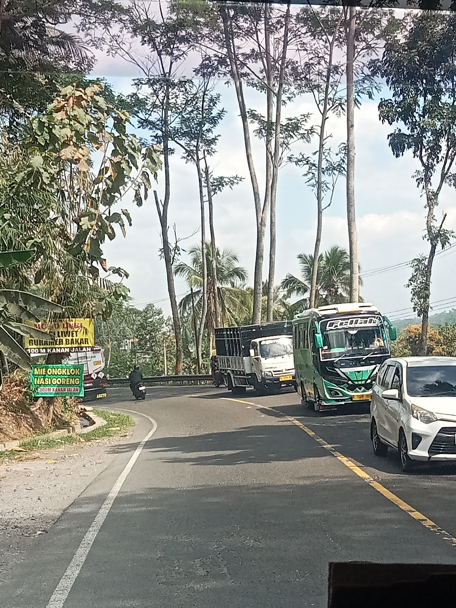 The image captures a street scene in Selokromo, Indonesia, with a winding asphalt road bordered by vegetation and tall trees. A white car is in the foreground, facing right. Behind it, a green and white bus with "Fellah" written on the windshield is traveling in the same direction, followed by a white truck. A motorcycle with a rider is in front of the truck. Two green banners are affixed to the left side of the road. The banner on top reads "MUSIM UDANG SELIWET GURAMEH BAKAR 100 M KANAN JALAN." The banner below it says "MIE ONGKLOK NASI GORENG 300M KANAN JALAN." The sky is visible through the trees.