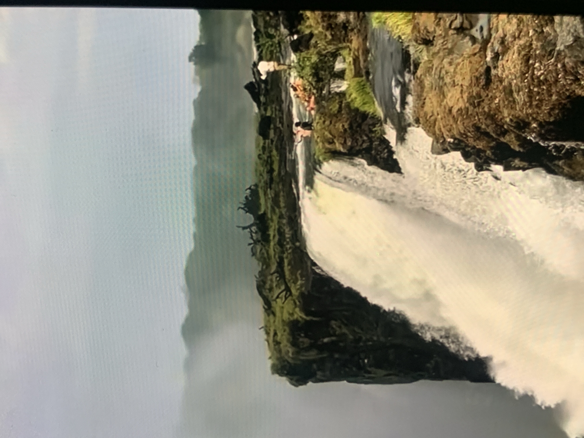 This is a vertical, medium-distance, eye-level shot of a wide waterfall. The water crashes over a sheer cliff face covered in moss and rocks. The cliff is lined with green vegetation and some dead branches. On the edge of the cliff, several people are visible. One person stands near the edge, looking out, wearing a light-colored shirt and dark pants. A group of people are seated or lying down further back, some in orange clothing. The background shows a hazy landscape of rolling hills covered in greenery, with a pale sky above. The overall impression is of a natural, rugged landscape with human presence at the precipice of a powerful waterfall. This image is not located in Stavropol, Russia.