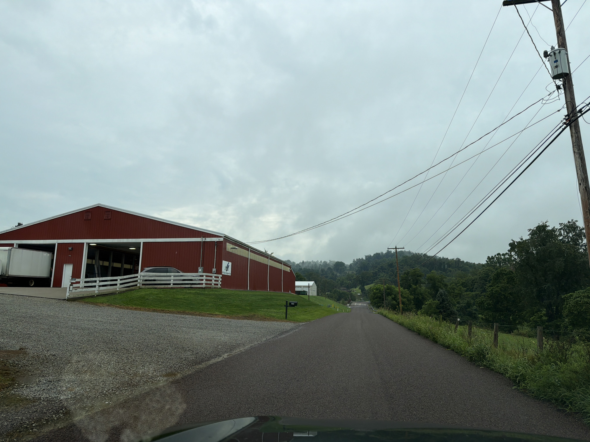 The image depicts a rural scene in Fallowfield Township, United States of America, on a cloudy day. A large red barn with white trim is visible on the left side of the frame, with a white trailer parked at its entrance. A white fence runs along the front of the barn, separating it from a grassy area. A paved road leads away from the barn and into a wooded, hilly landscape. A wooden utility pole with a transformer stands on the right side of the road, with power lines stretching across the sky. The overall atmosphere is overcast and serene, with no people visible in the frame.