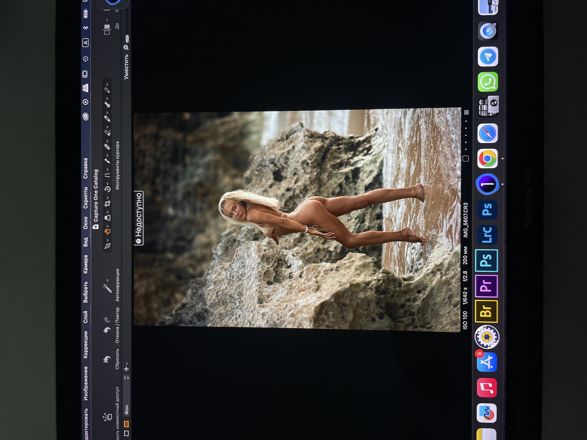 The image shows a screen capture of a Macbook Pro, with an open application displaying a photograph of a woman posing on a rocky shoreline. The woman, who appears to be blonde, is lying on her stomach with part of her body in the water. She is wearing a light-colored bikini. The background consists of rock formations and the sea. Icons visible in the Macbook's dock include Adobe Bridge, Adobe Premiere Pro, Photoshop, Lightroom, Capture One, and other macOS apps. Software application menus and toolbars are at the top of the open photograph. The image file name is IMG_5607.CR3. The EXIF data indicates ISO 100, 1/640 second exposure, f/2.8 aperture and 200mm focal length. There is no indication of Kazan, Russia in the image.