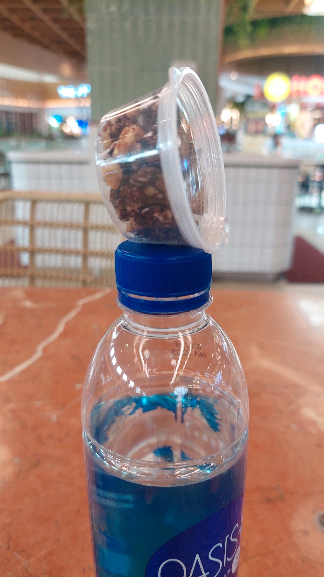 In a well-lit indoor environment, likely a mall food court in North Jakarta, Indonesia, a plastic bottle of Oasis brand blue beverage is upright on an orange-patterned table. The bottle's cap is a vibrant blue. Perched precariously atop the blue cap is a small, clear plastic container, overturned and filled with a mixture of granola and nuts. The background is blurred, but suggests a public space with seating and commercial establishments. The focus is on the unusual arrangement of the food container on the water bottle.