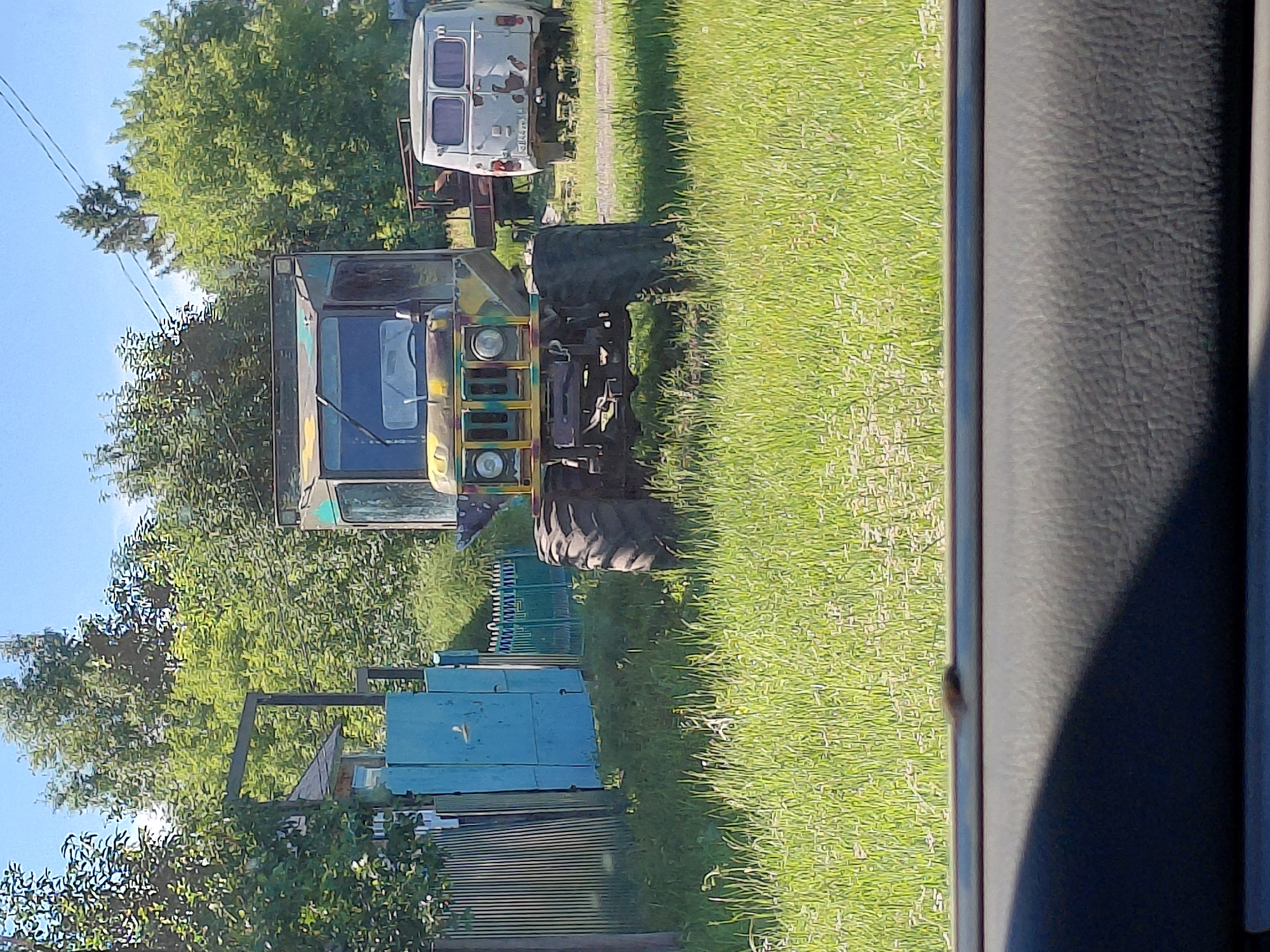 The image depicts a daytime outdoor scene in Novoselovo, Russia. The primary focus is a vintage, camouflaged military-style truck with large tires parked on a grassy area. Behind the truck, a white van is also visible. To the left, a blue gate and a weathered fence are present, along with trees and foliage. The foreground shows the interior of a vehicle, with a dark textured surface. The sky is clear and blue.