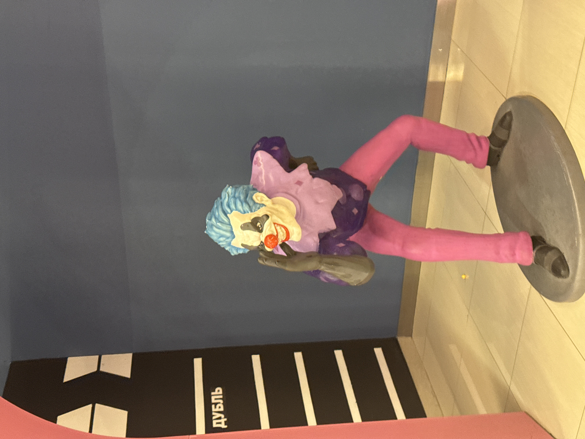 Here is a description of the image:
The image shows a Joker statue in the city of Perm, Russia. The statue features a clown with blue hair, white face paint, and a red nose. The clown is wearing a purple and dark purple outfit with pink leggings. The statue is placed on a gray circular platform. Behind the statue is a black and white striped object and some Russian text that reads "дубль", which translates to "double" in English. The statue is set against a wall that is divided into a blue top and a yellow/tan bottom.