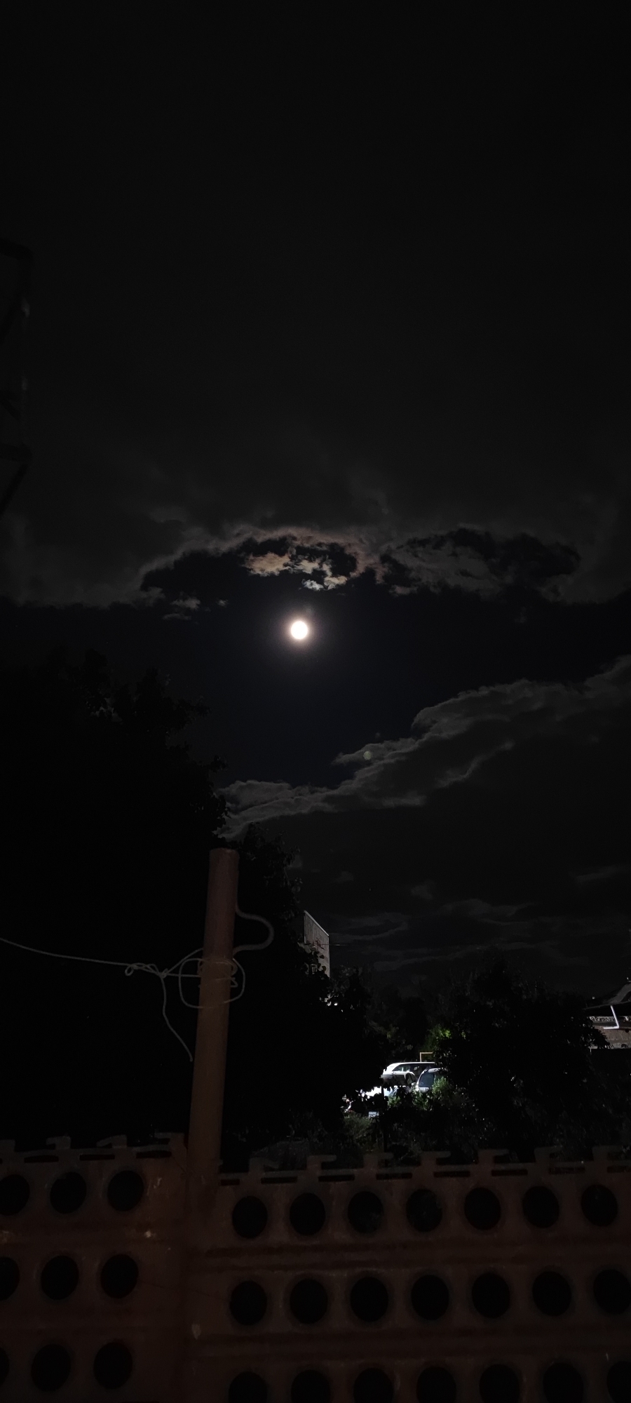 The image depicts a night scene in Yerevan, Armenia. A full moon is visible in the sky, partially obscured by clouds. Below the moon, there are trees and a building silhouetted against the dark sky. At the bottom of the image, there is a wall with circular openings. A pole with wires is seen in the foreground. In the distance, cars are parked.