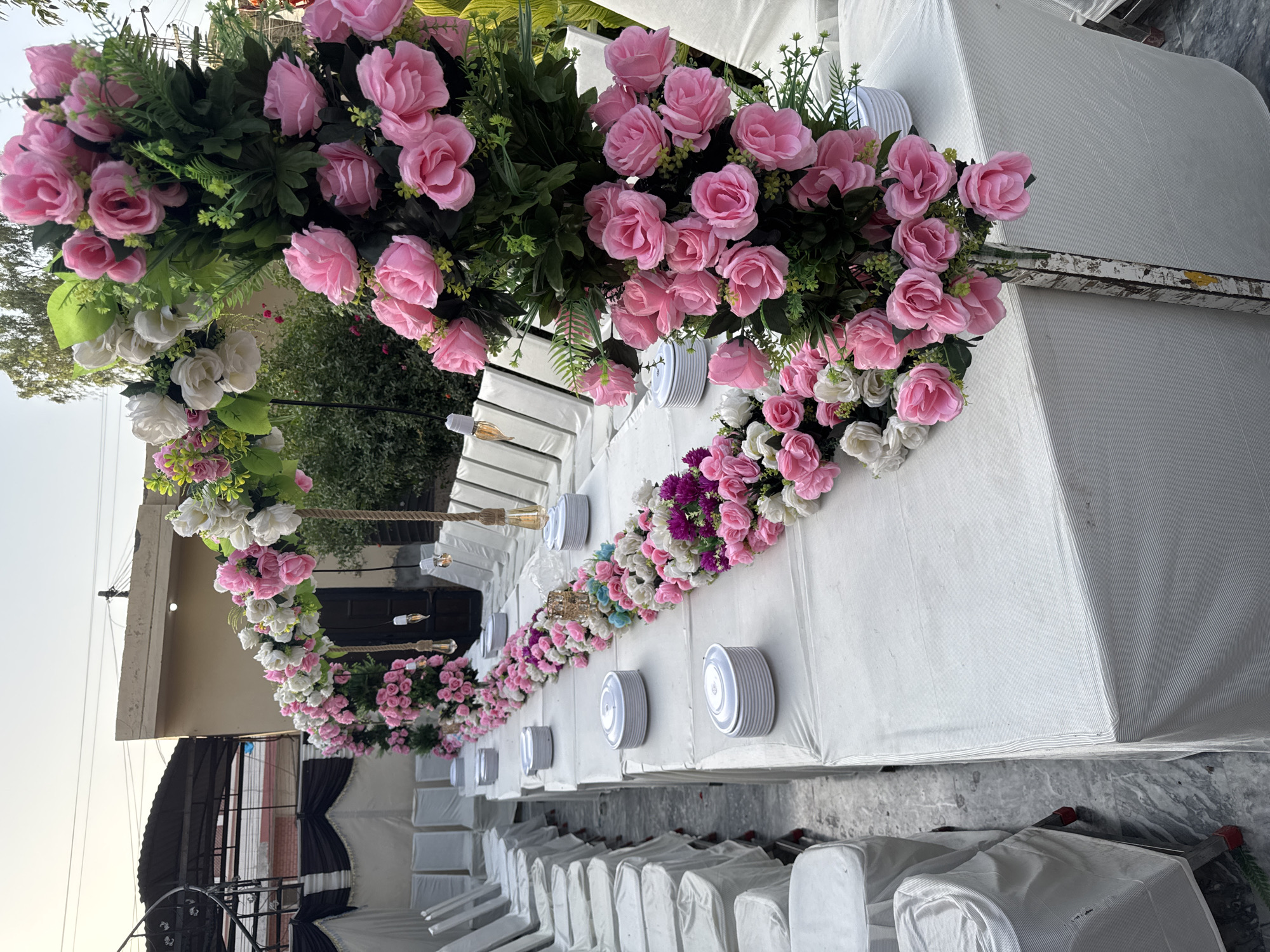 A long table decorated with pink and white flowers is set up outdoors. Plates are evenly spaced along the table. Rows of white chairs face the table on one side. The location is Justice Colony, Pakistan.