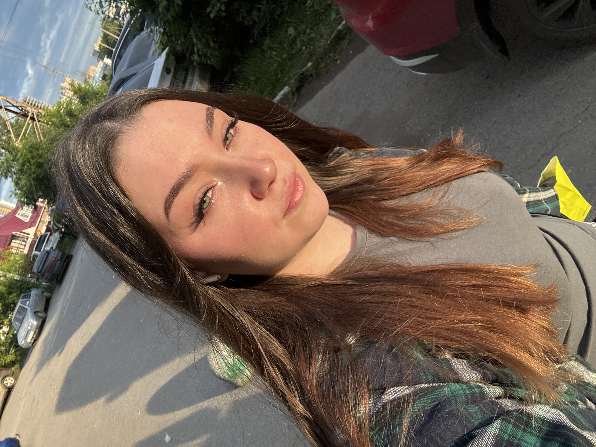 A selfie taken from a high angle shows a young woman standing outdoors on a paved surface, likely a street or parking lot, with a row of parked cars in the background. The woman has long, dark brown hair, green eyes, and is wearing a grey t-shirt and a green and blue plaid shirt. She has on wireless earbuds. The lighting suggests it is late afternoon or early evening, with long shadows cast across the ground. Behind her, several cars are visible, including a silver sedan and a dark-colored SUV. In the distance, buildings and trees can be seen, along with a cloudy sky. The presence of buildings, vehicles, and paved infrastructure indicates an urban or suburban setting. Based on the style of the buildings and cars, this scene is consistent with a location in Perm, Russia.