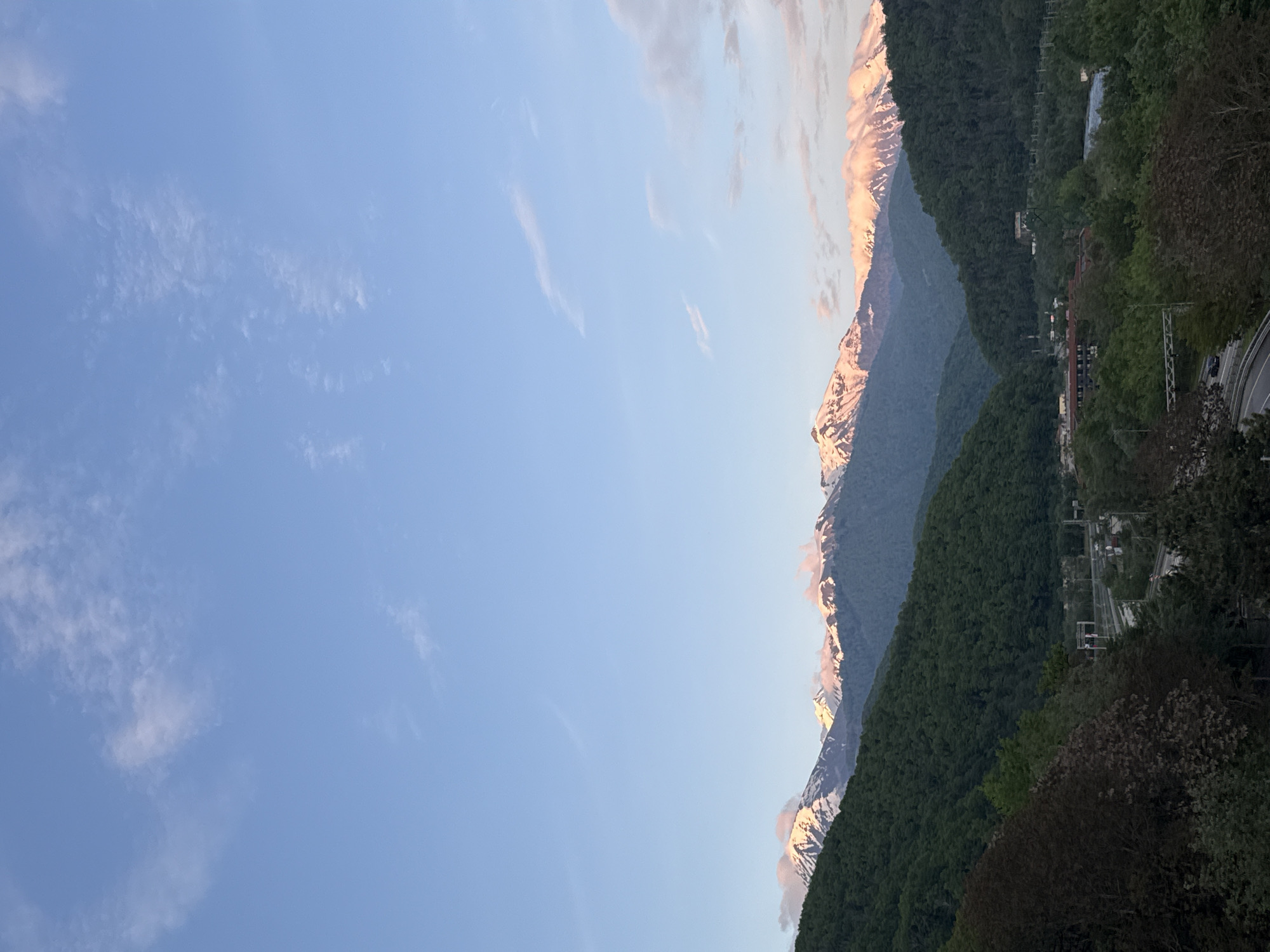 The image captures a mountainous landscape in Krasnaya Polyana, Russia. Snow-capped peaks are visible, with the sun catching their tops. Lush green forest covers the lower slopes of the mountains. A clear blue sky with some wispy clouds occupies a significant portion of the image. A winding road is partially visible among the trees, and there are structures that might be buildings or infrastructure related to the ski resort in the area.