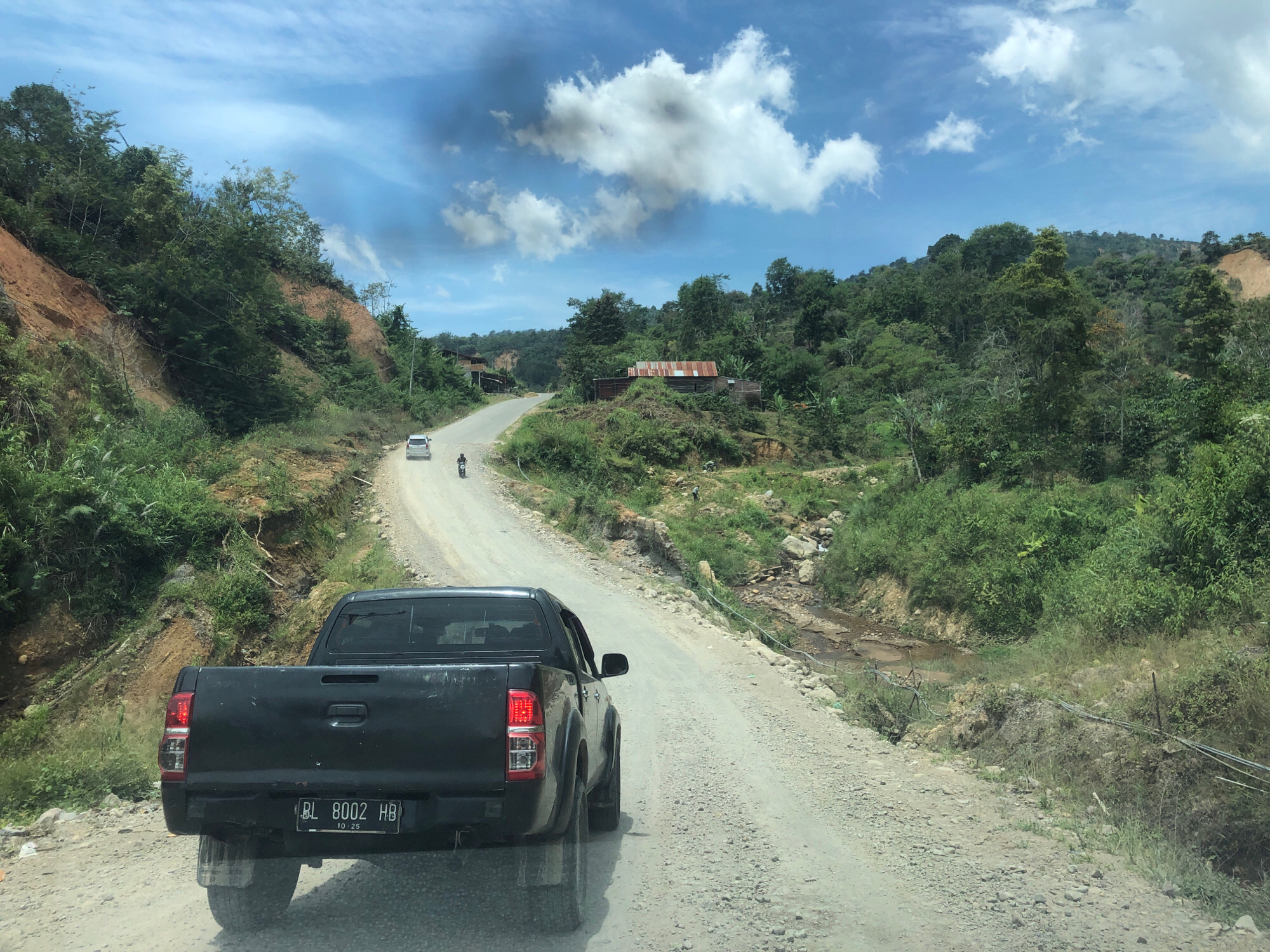 A black pickup truck with license plate BL 8002 HB is driving on a dirt road through a rural, mountainous area. The road is winding and appears to be under construction or in disrepair, with exposed dirt and rocks. Green vegetation covers the hillsides, and a small stream runs alongside the road in some sections. In the distance, there are a few simple buildings with corrugated tin roofs, suggesting a small settlement. The sky is blue with scattered white clouds, indicating it is daytime. The scene appears to be in a remote location, possibly in Bintangpermata, Indonesia, given the details provided. There are no people clearly visible in the foreground, but a few distant figures and a motorcycle can be seen on the road ahead.