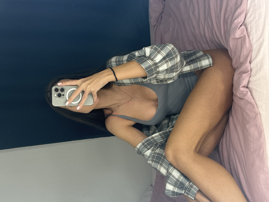 The scene depicts an indoor residential setting in Saint Petersburg, Russia. A single individual, presumed female, is seated on a bed covered by a light pink duvet. The subject is wearing a grey bodysuit and an open, long-sleeved plaid shirt in grey and white tones. A thin gold-colored chain with a cross pendant is visible on the subject's neck. A dark, thin bracelet is present on the left wrist. The subject's face is obscured by a smartphone, identified as an iPhone with a clear case and a visible MagSafe ring, held by the right hand. A ring is worn on the right hand's ring finger, and the fingernails are painted a light, possibly pink, color. The background features a wall partitioned vertically into a light grey section on the left and a dark blue section on the right.