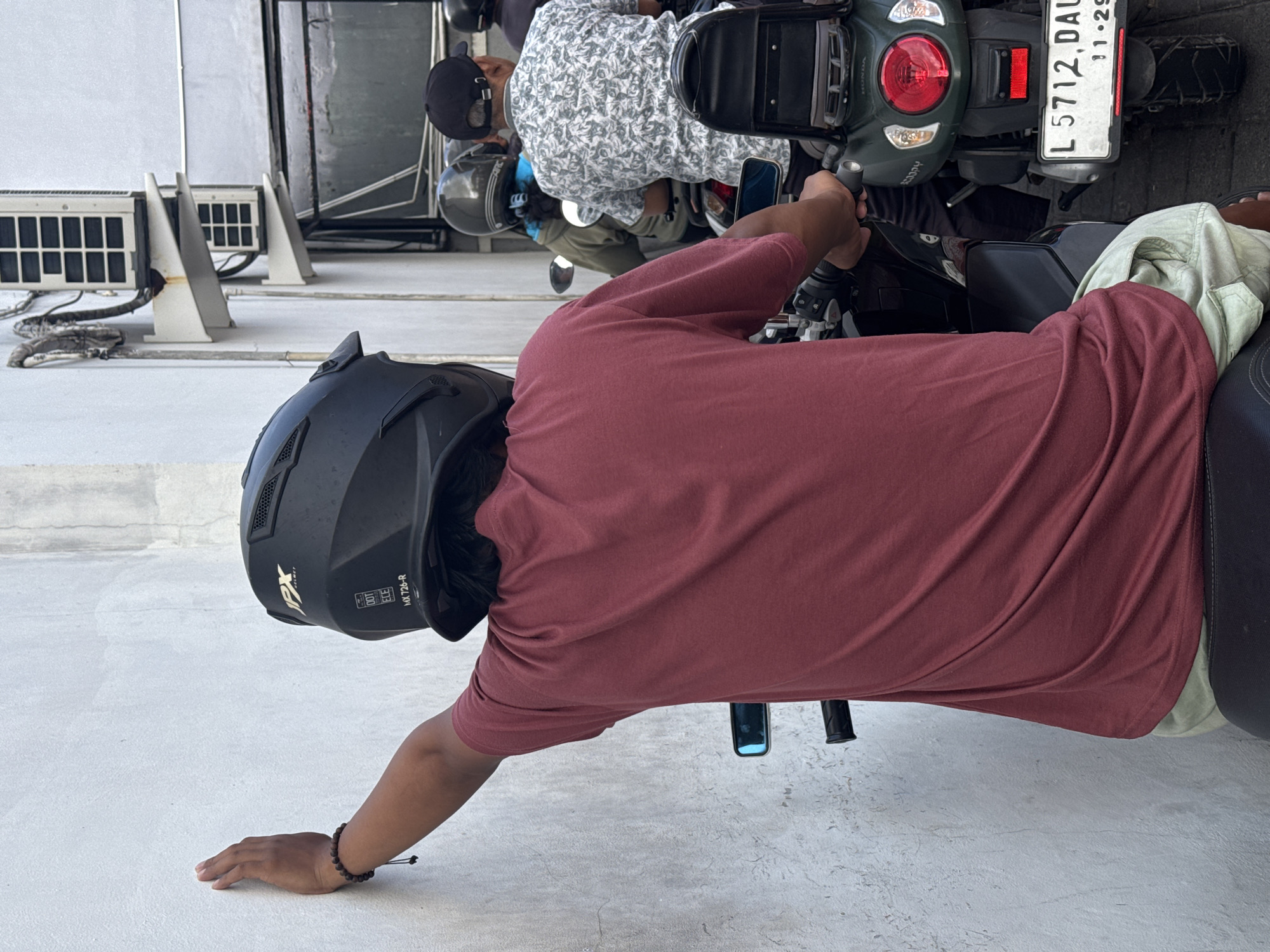 A ground-level medium shot depicts a man on a motorcycle on a concrete surface. The man is wearing a maroon t-shirt, khaki shorts, and a black helmet. He is leaning forward, with his left hand touching the ground. He wears a beaded bracelet. A motorcycle with the license plate L 5712 DAU is visible. Other people are riding motorcycles nearby. In the background, three outdoor air conditioning units are attached to the side of a building. The image was taken in North Kuta, Indonesia.