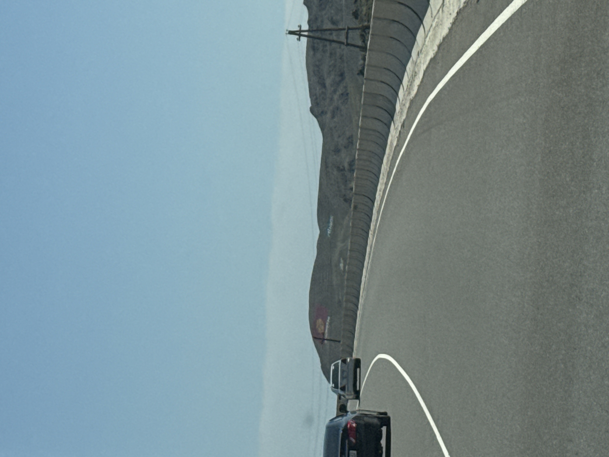 The image captures a view from a vehicle traveling on a paved road, likely a highway, in the vicinity of Kok Moynok Eki, Kyrgyzstan. The road curves gently to the right, marked by a white line on the asphalt. To the right of the road, a concrete barrier with a segmented design runs along the edge. Beyond the barrier, a barren, hilly landscape is visible under a clear blue sky. A single, tall wooden utility pole stands on the right side of the frame, supporting overhead wires. In the distance, on top of one of the hills, a red and yellow flag is mounted on a pole. In the foreground, several cars are on the road. A black sedan is in the left lane, moving away from the camera. A white sedan is in the right lane, also moving away. The overall scene depicts a rural or semi-rural environment on a bright, clear day.