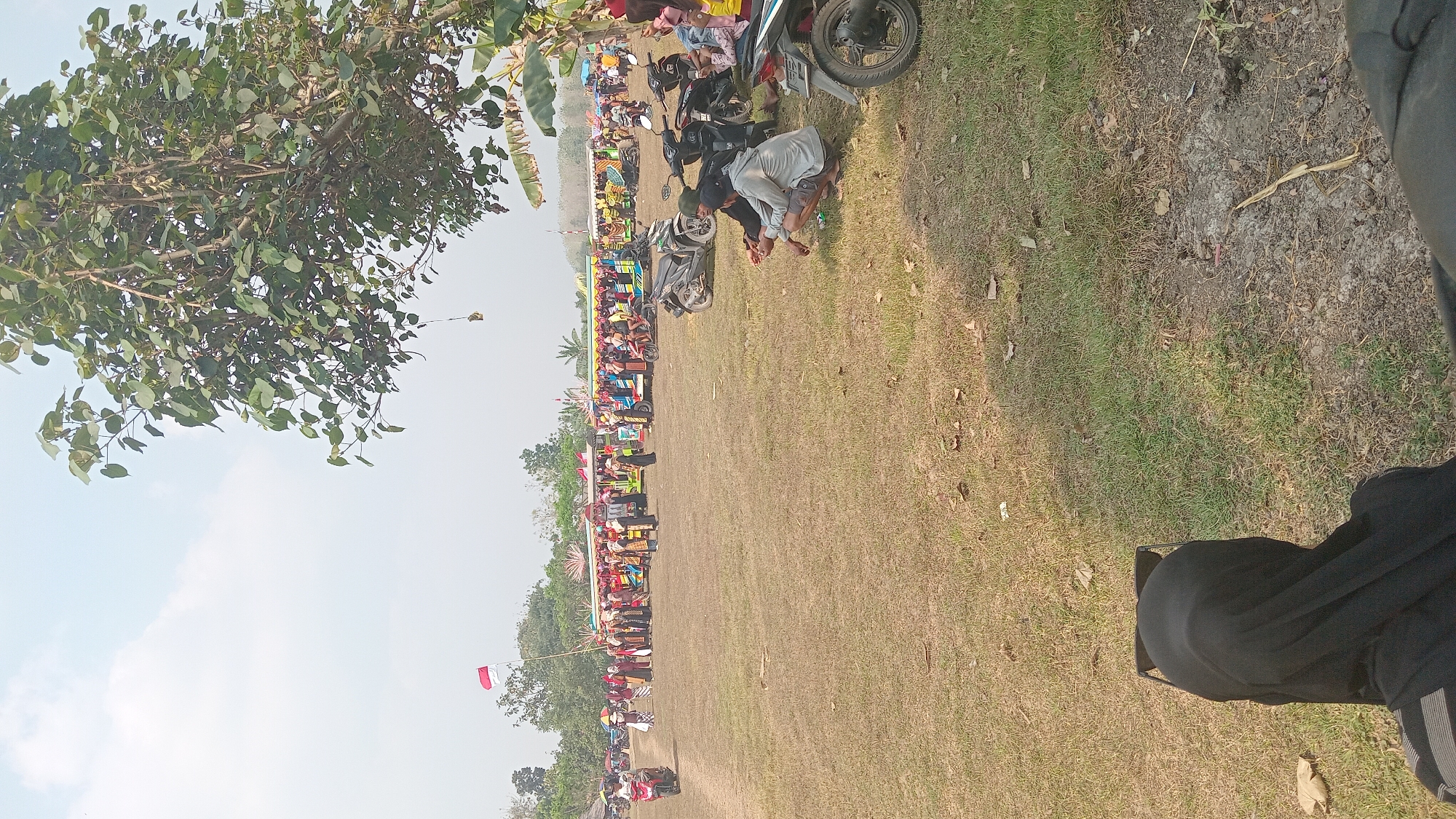 In Kedungwaru, Indonesia, an outdoor event is taking place on a dry, grassy field under a hazy sky. The scene is populated with several groups of people. In the foreground, a person wearing a black hijab and sunglasses is seated, facing away from the camera. Further into the field, a group of men are crouched down, possibly observing something or resting. Several motorcycles are parked in the middle ground, some of them occupied. In the background, a crowd of people is gathered around decorated vehicles, some of which appear to be small trucks or buses. An Indonesian flag flies prominently on a flagpole among the assembled group. The environment is sparsely vegetated, with a large tree with lush green leaves dominating the right side of the frame and some smaller trees and structures visible in the distance. The overall impression is of a local gathering or celebration in a rural or semi-rural Indonesian setting.