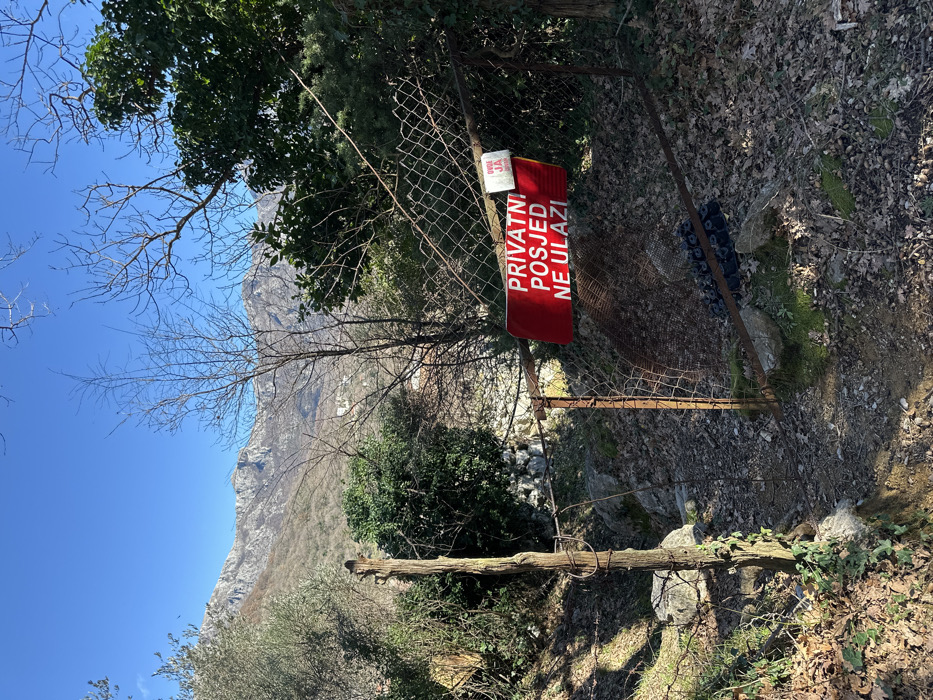 This image presents an outdoor scene in the Zupci area of Montenegro, featuring a path leading to a property boundary under a clear blue sky. The foreground shows an unpaved dirt path covered with scattered dry leaves and small rocks. To the immediate left, a weathered, truncated wooden post anchors a section of wire mesh fence. The central feature is a rusted, wire mesh fence, approximately waist-height, with an opening that appears to be a former gate. Attached prominently to this fence is a rectangular red sign with white capital letters reading "PRIVATNI POSJED NE ULAZI," which translates to "PRIVATE PROPERTY DO NOT ENTER." A smaller, partially obscured white sign with red text, reading "OVDE JA..." (Here I...) is affixed above the main sign. Behind the fence, a collection of dark, possibly rubber, debris is visible on the ground. The midground is characterized by varied vegetation, including dense green shrubs and several bare deciduous trees, indicative of late autumn or winter. In the background, rugged, vegetated mountains rise under a stark, clear blue sky. Distant, light-colored structures, likely buildings, are discernible on the lower slopes of these mountains. No individuals are present, and no actions or interactions are depicted.