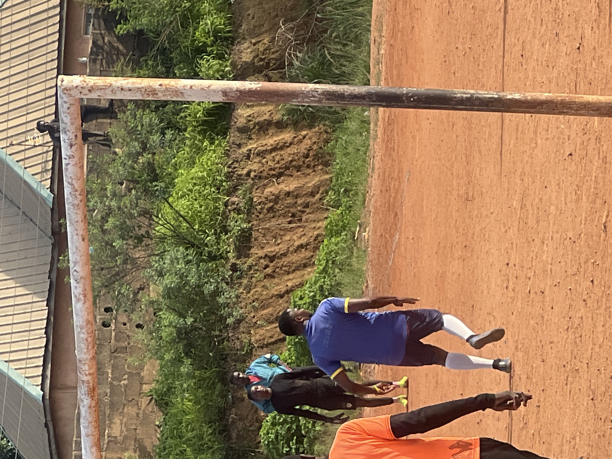 The image depicts a dusty, outdoor football field in Jos, Nigeria. In the foreground, a person in an orange shirt and black sleeves walks away from the camera, their back partially visible. To their right, a man in a blue shirt with yellow accents and dark shorts is in motion, facing away from the camera and towards the goalpost. He is wearing white socks and dark shoes.
Behind him, a person in a black long-sleeved shirt and black shorts stands with their hands on their hips, looking towards the viewer. Further back, another person in a light blue jacket over a red shirt stands with their hands in their pockets, also looking towards the viewer.
The football goalpost, with a rusty horizontal bar and a weathered vertical pole, is prominently positioned in the center of the frame. Beyond the field, a steep, earthen embankment covered in green vegetation rises. In the background, there is a building with a corrugated metal roof and a brick wall. A person can be seen standing on top of the brick wall, looking down at the scene. The overall impression is that of a casual football game or training session taking place on a typical unpaved field.