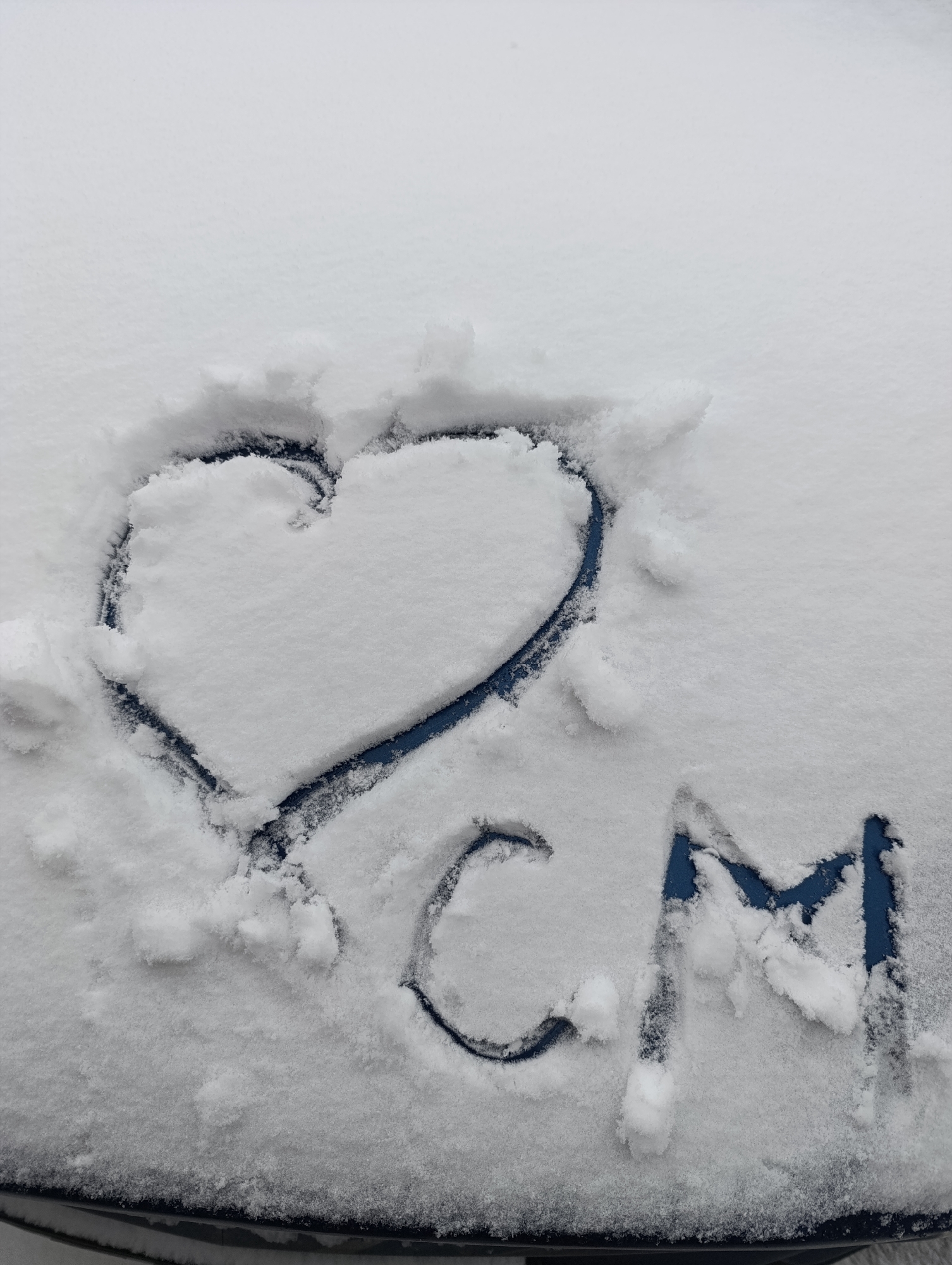 The image shows a heart and the letters "CM" drawn in the snow on a blue surface, likely a car hood. The scene appears to be outdoors, with a layer of fresh snow covering the surface. The snow is white, and the markings reveal the blue color of the car underneath.