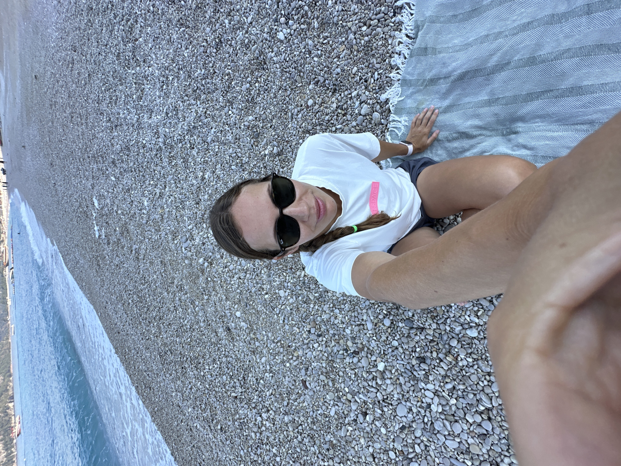 A woman is sitting on a pebble beach in Fethiye, Turkey, with the sea to her left. She is wearing a white t-shirt with a pink graphic on it, sunglasses, and dark shorts. She has a braid in her hair and is wearing a smartwatch on her left wrist. She is sitting on a striped blanket. The sea is blue and calm, with a few boats visible in the distance. The beach is covered in small, grey pebbles. The sky is overcast.