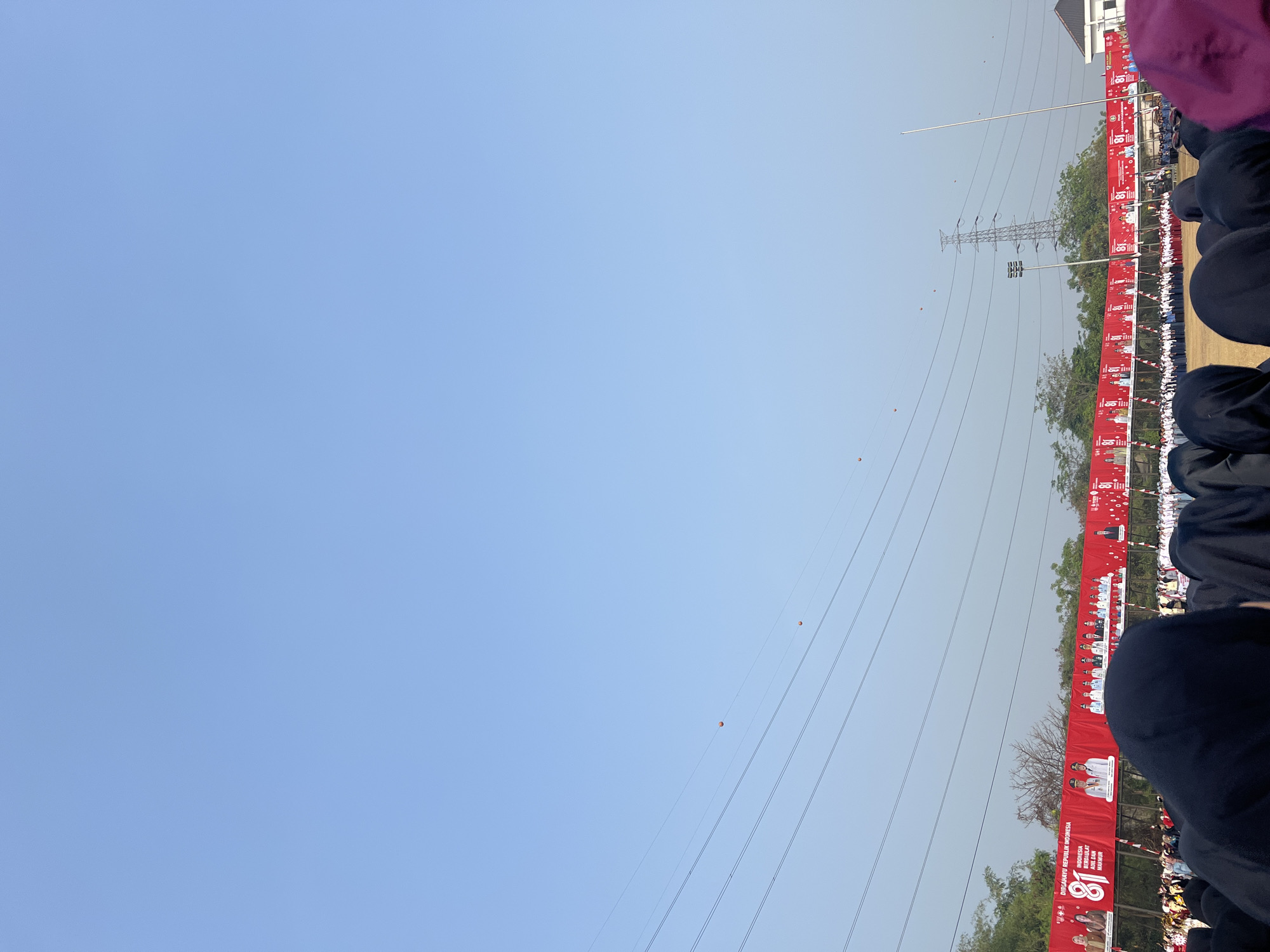 The image depicts an outdoor event with a clear blue sky and several power lines crisscrossing the upper portion of the frame. A large red banner stretches across the middle ground, featuring text that reads "DIRGAHAYU REPUBLIK INDONESIA 81" and portraits of various individuals. Below the banner, a crowd of people, many wearing head coverings, are gathered on a field of dry grass. The background shows trees, a tall transmission tower, and a building. The overall scene suggests a public celebration or gathering. The location is identified as Sukamantri, Indonesia.