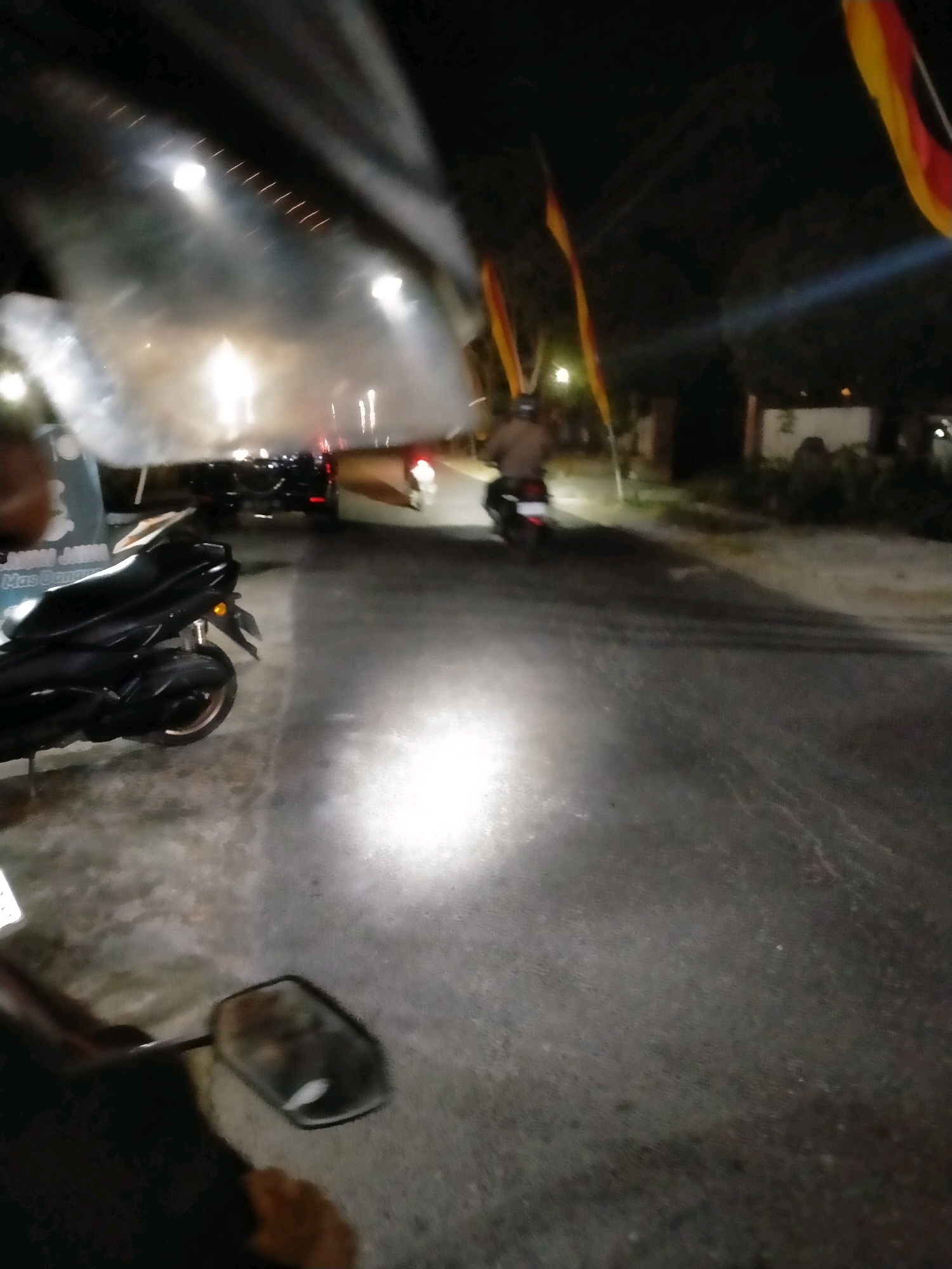 This is a low-light, blurry photograph taken at night in Playen, Indonesia, likely from the perspective of someone riding a motorcycle. A black motorcycle is prominently featured in the left foreground. In the background, several other vehicles are visible, including cars and motorcycles, suggesting a street or road setting with traffic. Two flags with red and yellow stripes are visible in the upper right and left portions of the image, indicating a possible event or celebration. The road surface appears wet or reflective, casting a strong light from a source in the foreground. The overall atmosphere is dark and somewhat chaotic due to the motion blur.