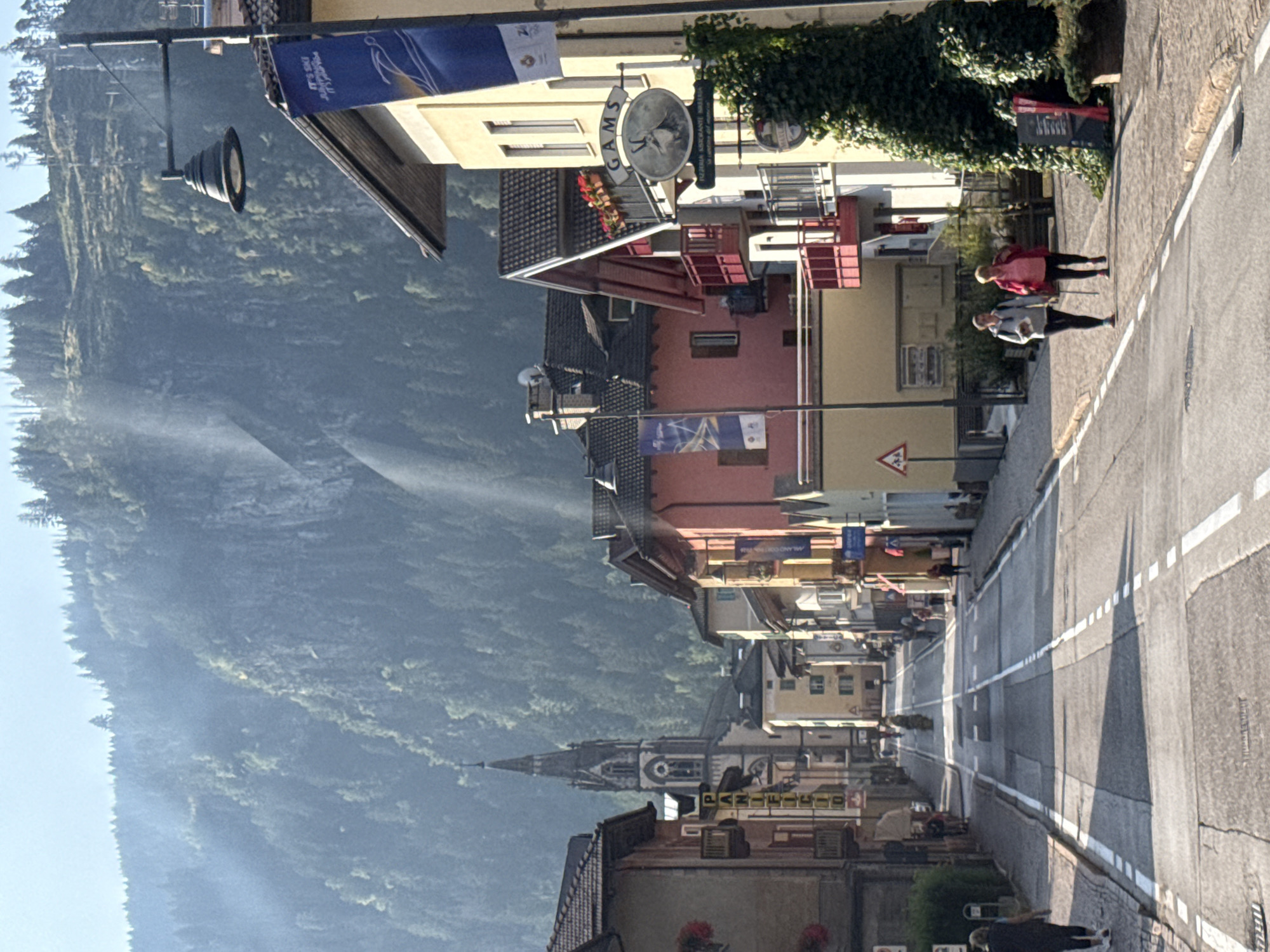 A sunlit street in the city of Predazzo, Italy, is lined with colorful buildings on either side. A prominent church spire rises in the background, partially obscured by trees. The street is paved and marked with white lines. Two women are walking on the sidewalk on the right side of the street. Several banners hang from lampposts, and a sign for "GAMS Pizzeria Ristorante Galleria" is visible on one building. The environment appears to be mountainous, with a heavily wooded slope forming the backdrop. The overall scene suggests a peaceful, daytime setting in a town nestled within a natural landscape.