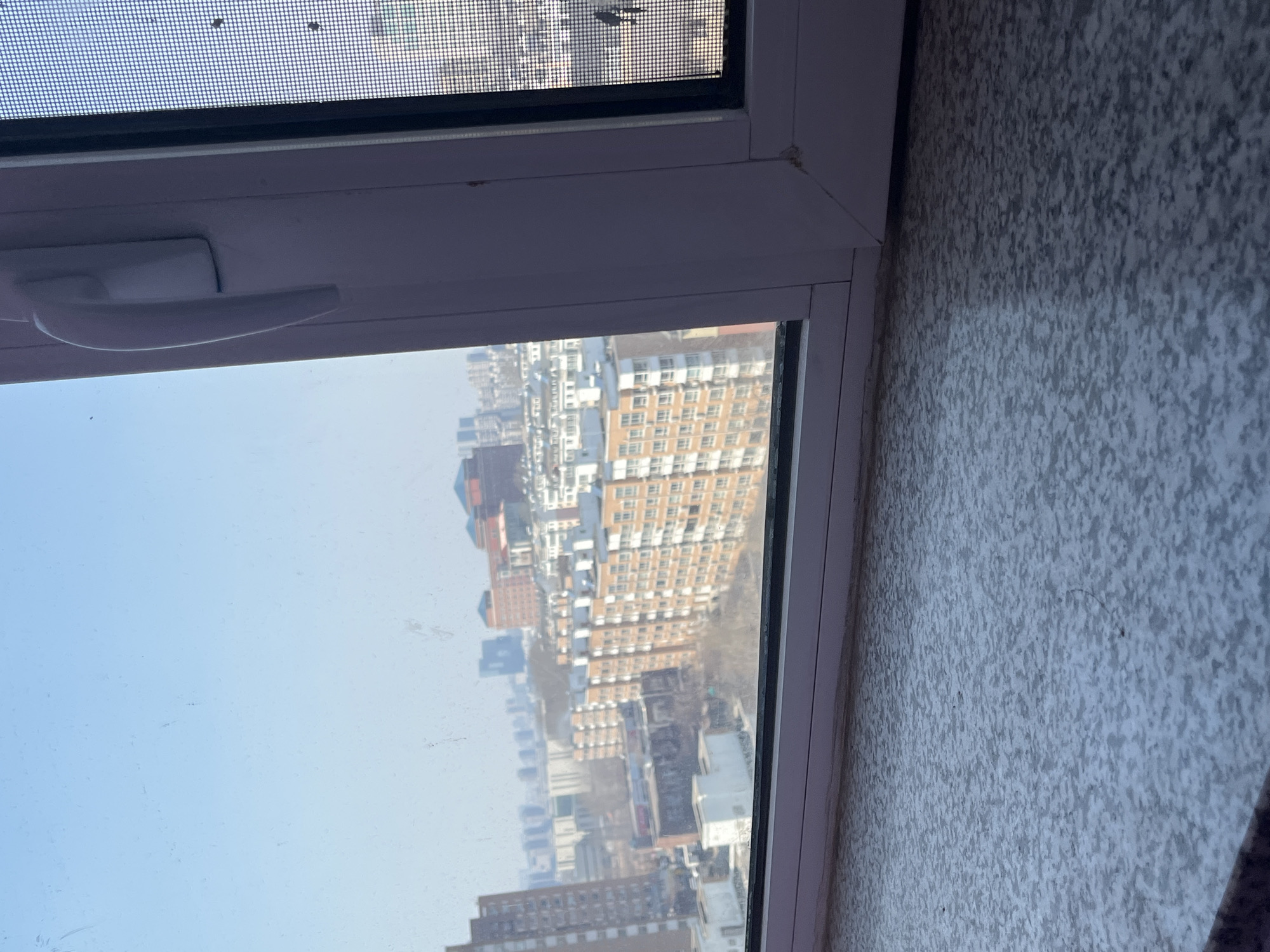 The image presents an eye-level view from an elevated interior position, looking out over an urban landscape in Changchun City, China, on a clear day. The immediate foreground features a grey, speckled window sill with visible dust and debris. Above the sill, a white window frame with a multi-pane structure is visible. The right-hand pane of the window is partially obscured by a dark mesh screen, while the central white vertical frame section includes a white, elongated handle positioned vertically.
Through the clear window glass, a dense cityscape extends into the distance under a light blue sky. The midground is dominated by numerous multi-story residential buildings, primarily displaying orange-brown facades with white window frames and balconies, arranged in blocks. Interspersed among these are some darker, possibly industrial or commercial structures. Sparse, leafless trees are visible between the buildings, indicating a season outside of full foliage. The background features a hazy skyline of taller, more modern high-rise buildings. The overall scene is devoid of any visible individuals or explicit activities. The window glass itself exhibits smudges and dust particles.