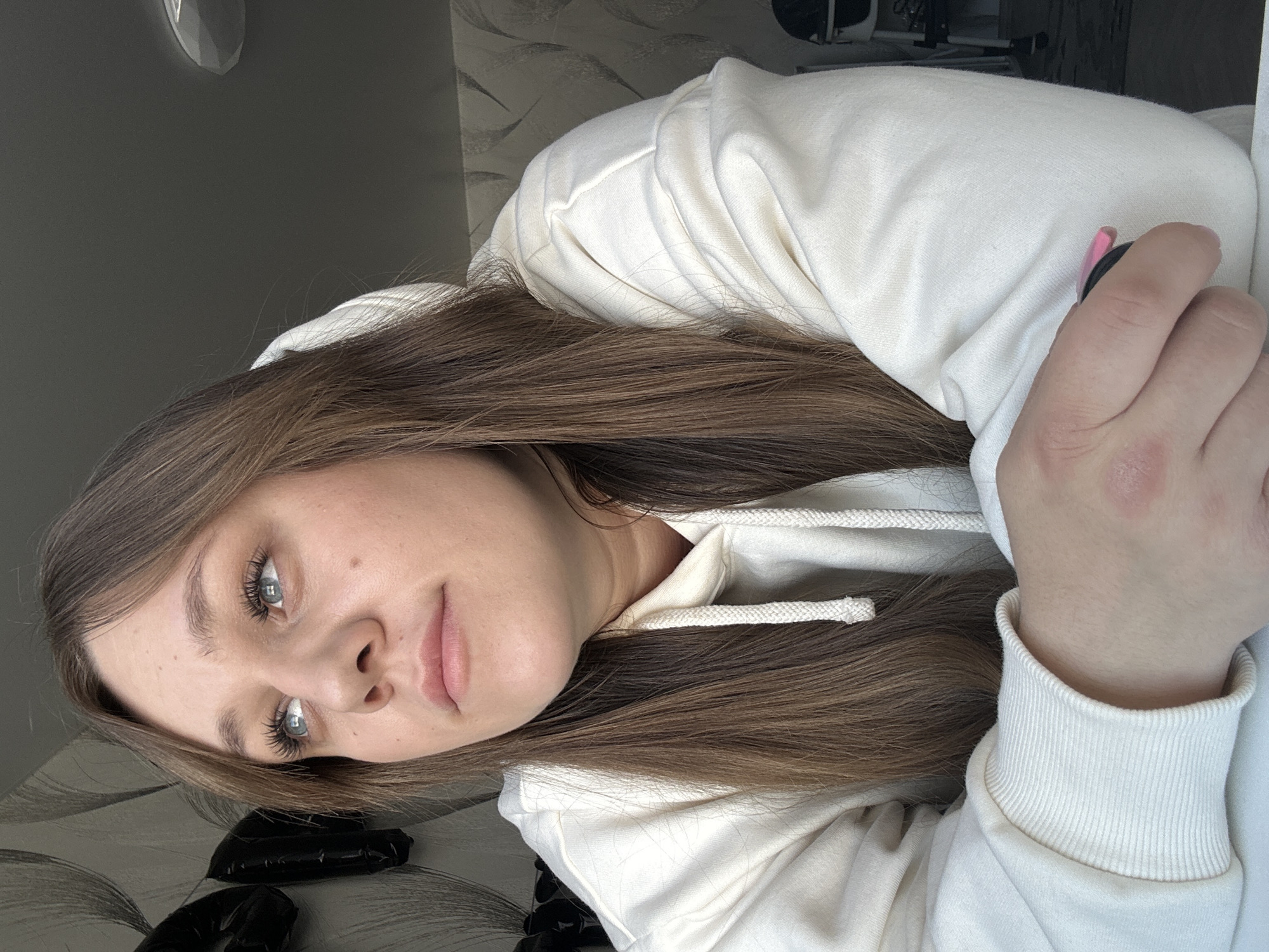 A close-up indoor image shows a woman with long, straight, brown hair and blue eyes looking upwards and to her left. She is wearing a cream-colored hooded sweatshirt with visible drawstrings. Her right forearm and hand rest on a light-colored surface. She holds a small, dark object with a pink accent in her right hand; a minor red abrasion is visible on the back of her right hand. The background consists of a wall covered in light-toned wallpaper featuring a large, abstract, dark grey swirling pattern. A black, inflated object is partially visible on the far left. The ceiling is light grey, with a circular light fixture partially visible on the upper right. Blurred elements in the far right background include a part of a high chair and a patterned floor covering. The specific location within the city of Chaikovsky, Russia, cannot be determined from visual evidence.