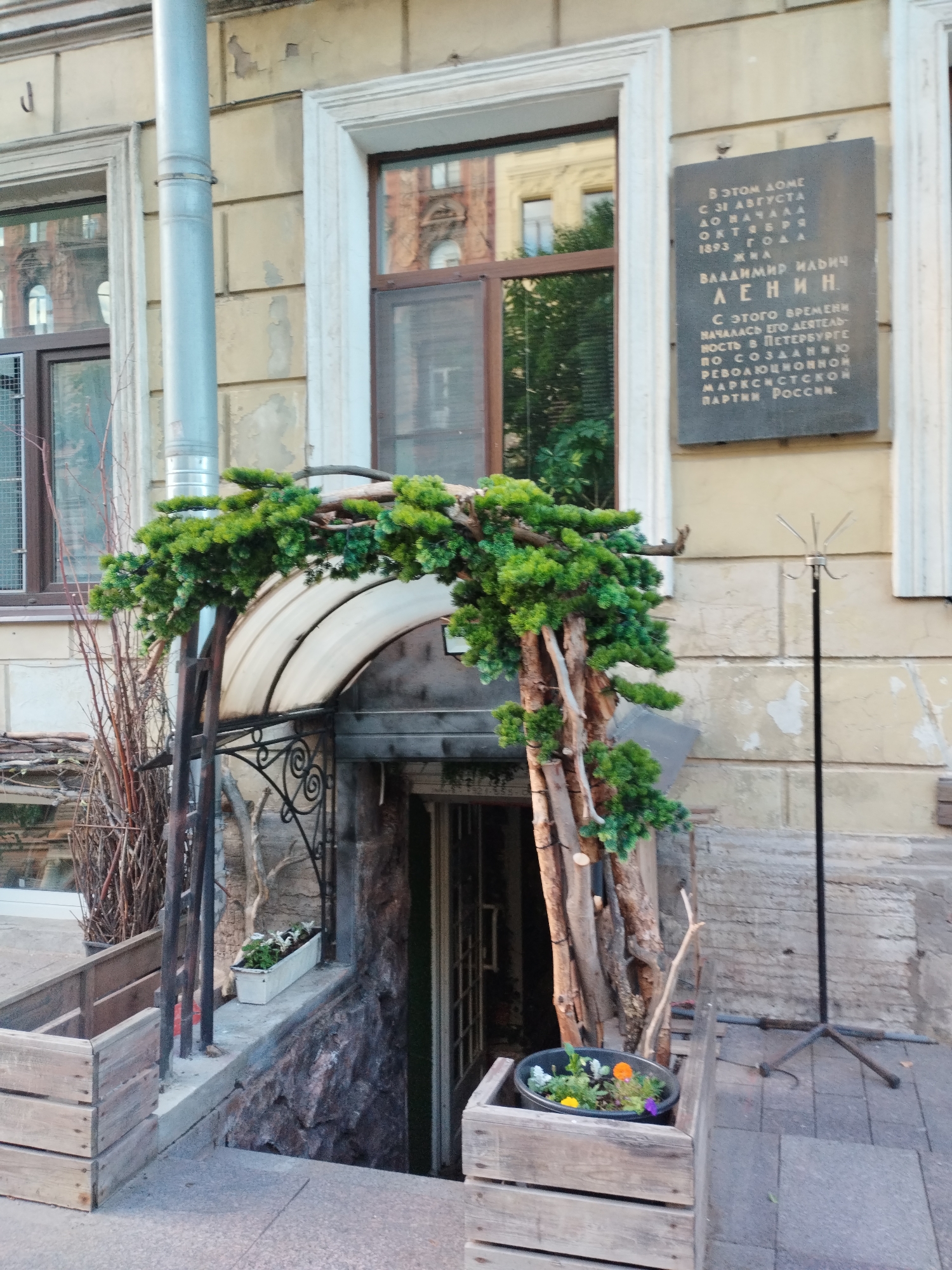 The image shows the entrance to a building with a memorial plaque. The plaque, mounted on a light yellow, weathered facade, is dark and rectangular, inscribed with Cyrillic text. It reads, "В ЭТОМ ДОМЕ С 31 АВГУСТА ДО НАЧАЛА ОКТЯБРЯ 1893 ГОДА НАЧАЛАСЬ ЕГО ДЕЯТЕЛЬНОСТЬ В ПЕТЕРБУРГЕ ПО СОЗДАНИЮ МАРКСИСТСКОЙ РАБОЧЕЙ ПАРТИИ РОССИИ. ВЛАДИМИР ИЛЬИЧ ЛЕНИН". This translates to, "In this house from August 31 to early October 1893, his activity began in Petersburg to create the Marxist Workers' Party of Russia. Vladimir Ilyich Lenin." The entrance itself is partially obscured by an archway decorated with evergreen branches and rustic wooden elements. Beneath the arch is a dark opening, suggesting a basement or lower-level entrance, with stone walls visible. To the left of the entrance, there are wooden crates and a metal pipe. To the right, a coat rack stands on a paved area. The overall scene suggests a historical site in Saint Petersburg, Russia, commemorating Vladimir Lenin's revolutionary activities.