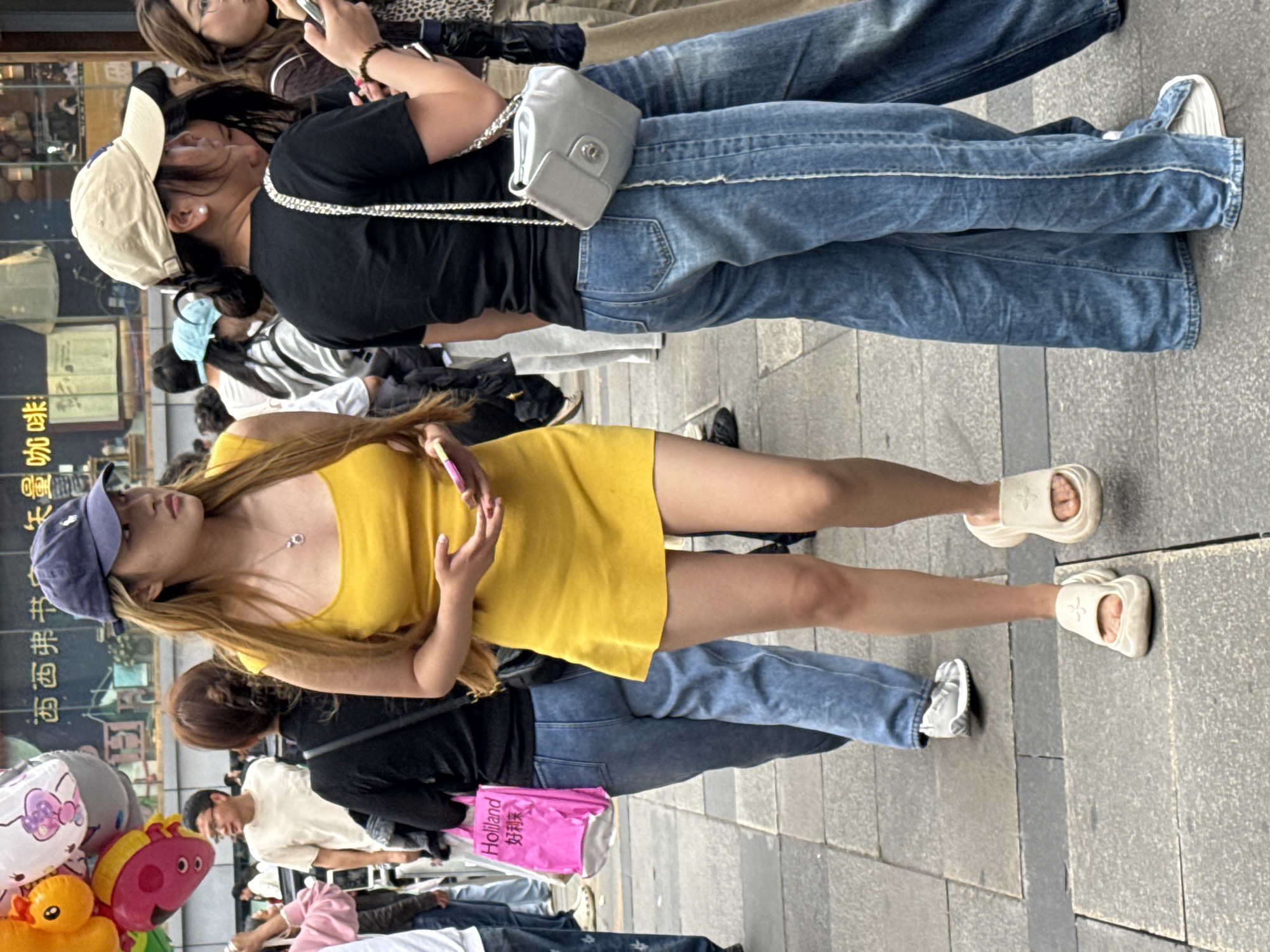 The image captures a street scene in Liangjiang, China. The foreground features a young woman wearing a yellow mini dress and a blue baseball cap. She is holding a smartphone. Behind her is a group of people, including a woman in a black shirt and jeans with a silver purse. There is a shopping bag with writing in Chinese that reads "Holland 好利来". Additional people and street decorations are visible in the background. The ground is paved with light gray tiles.