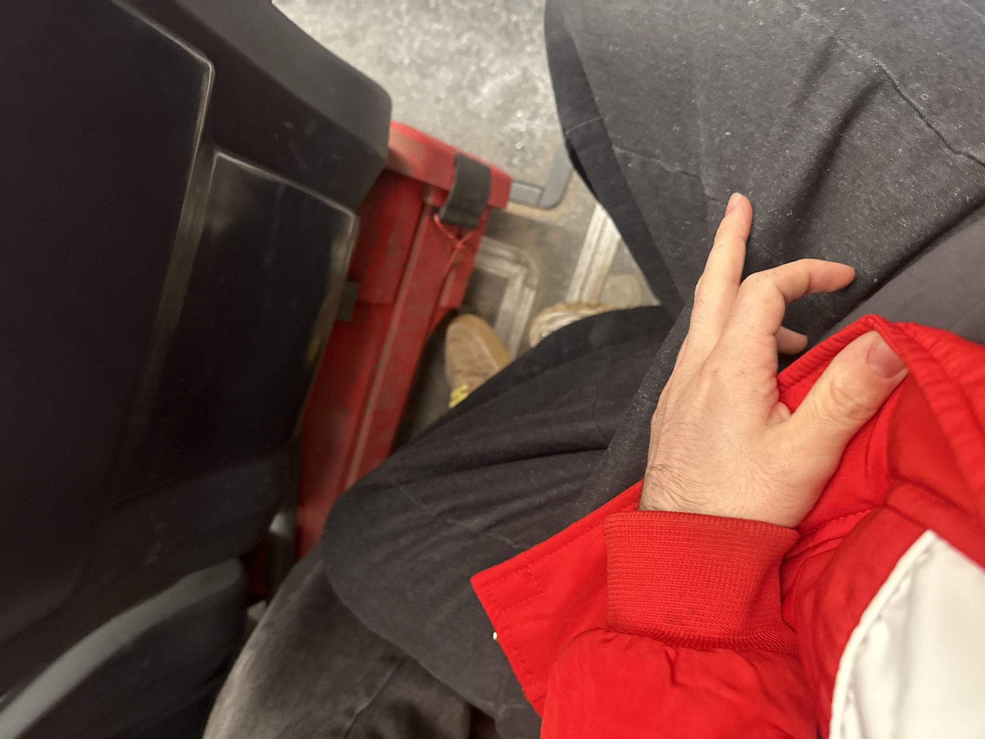 The image presents a close-up, overhead perspective. The scene includes a person seated, wearing dark gray pants and a red and white jacket. Their hand rests on the jacket. Adjacent to the person is a black object and a red container. Part of a tan shoe is visible. The setting implies an indoor location, possibly a vehicle, with a grey flooring or surface in the background. The image doesn't provide enough information to confirm the location as Khimki, Russia, or any specific city.