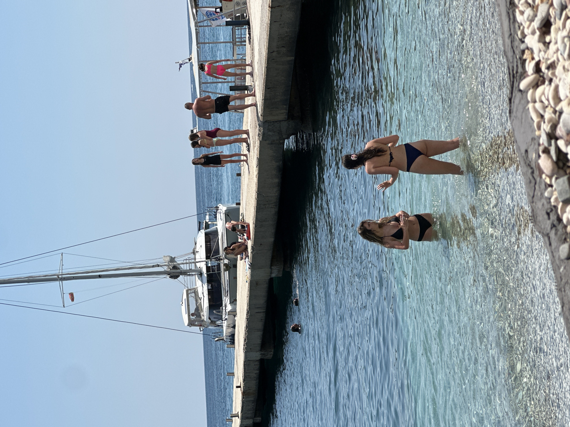 This is a vertical, eye-level shot of a sunny day at a pier in Budva, Montenegro. The scene depicts several people enjoying the clear, shallow water. On the pier, a group of individuals stands or sits near a docked boat. One person in a pink bikini is looking out at the water. Further down the pier, four people stand in a line, one in a black dress, another in a yellow bikini, and two in maroon bikinis. A man in black shorts stands in front of them. In the water, three women are visible. Two are in bikinis, one in black and another in dark blue. One woman is wading near the shore, while the other two are further out, with their heads visible above the water. The water is very clear, allowing the pebbled seabed to be seen. The weather appears to be warm and pleasant, indicated by the attire and activities of the people. The sun is high in the sky, suggesting it is likely midday.