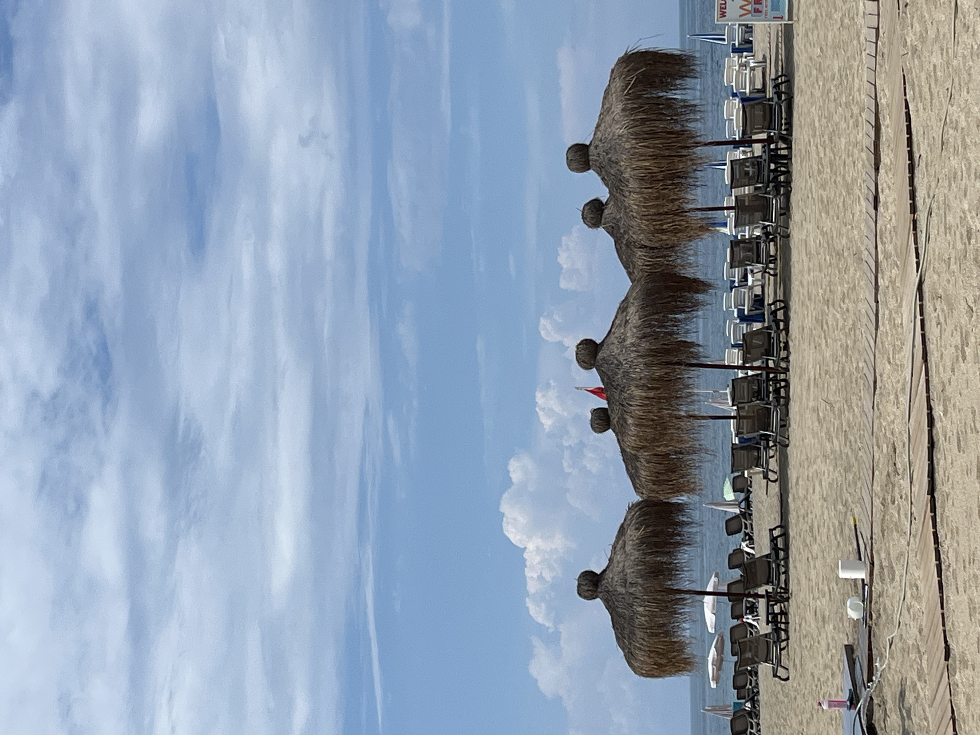The image shows a beach scene in Alanya, Turkey. Rows of straw parasols stand on the sandy beach, along with beach chairs underneath. The sky is blue with scattered clouds. A wooden walkway borders the beach. The sea is visible in the background.